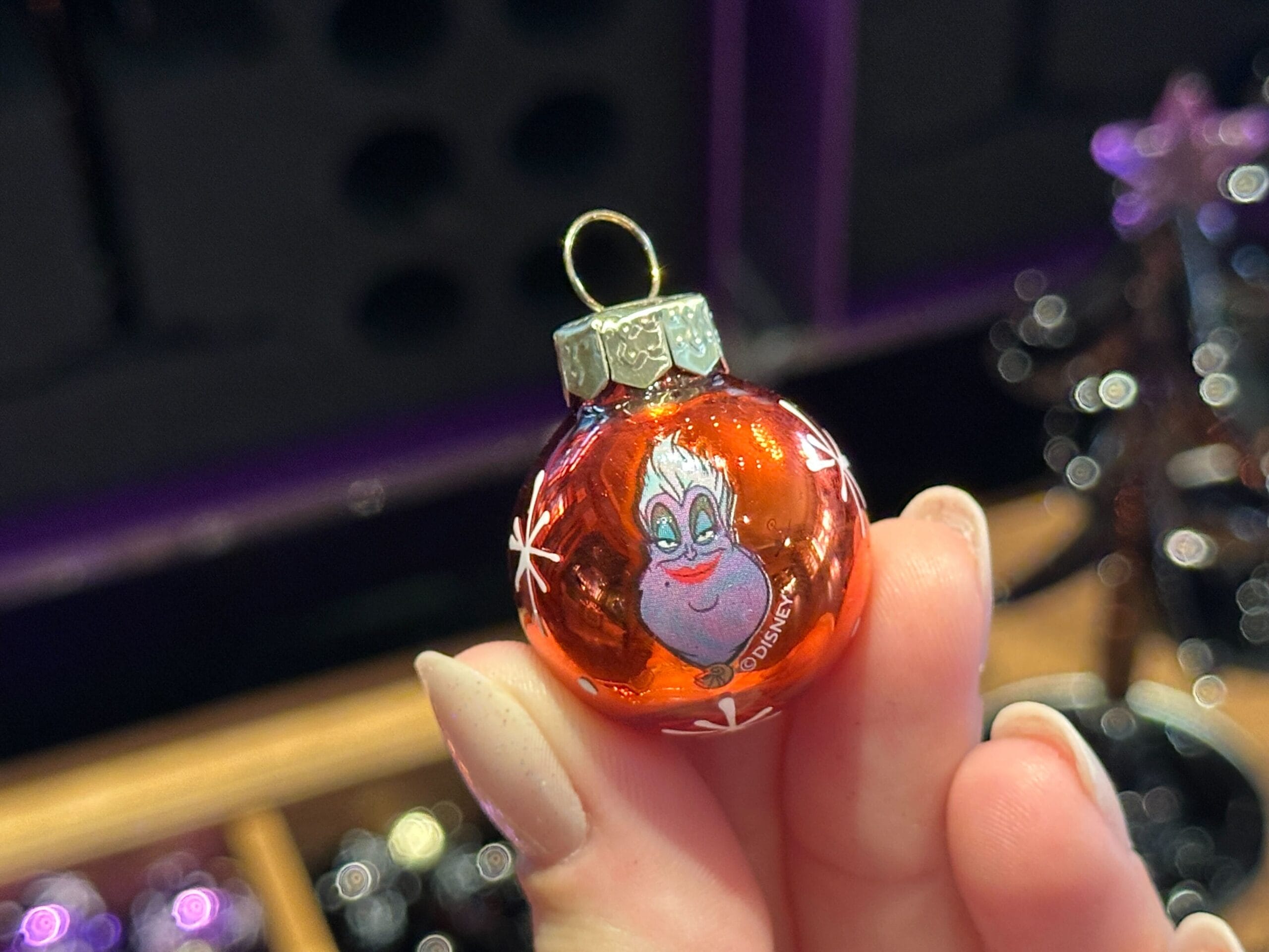 A hand holds a red glass Villain Ornament featuring Ursula from Disney’s The Little Mermaid, with a festive background.