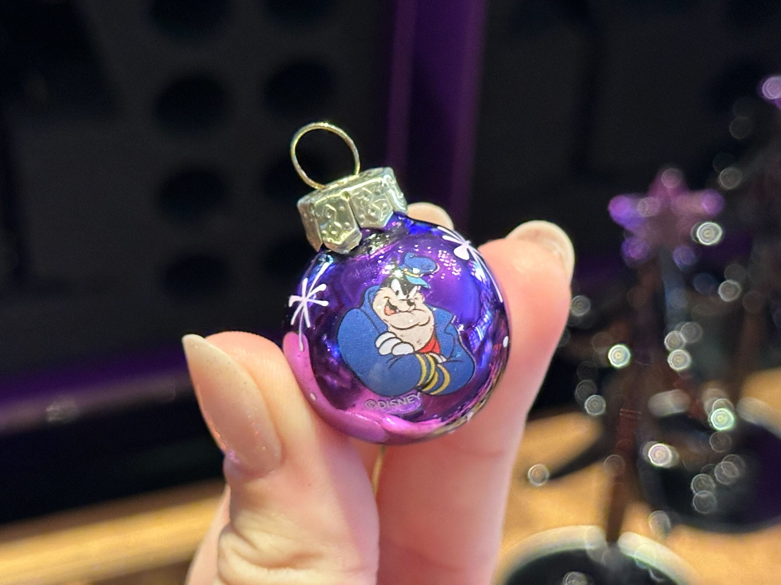 A hand holds a small purple Villain Ornament featuring Genie from Disney's "Aladdin" with a gold cap on top.