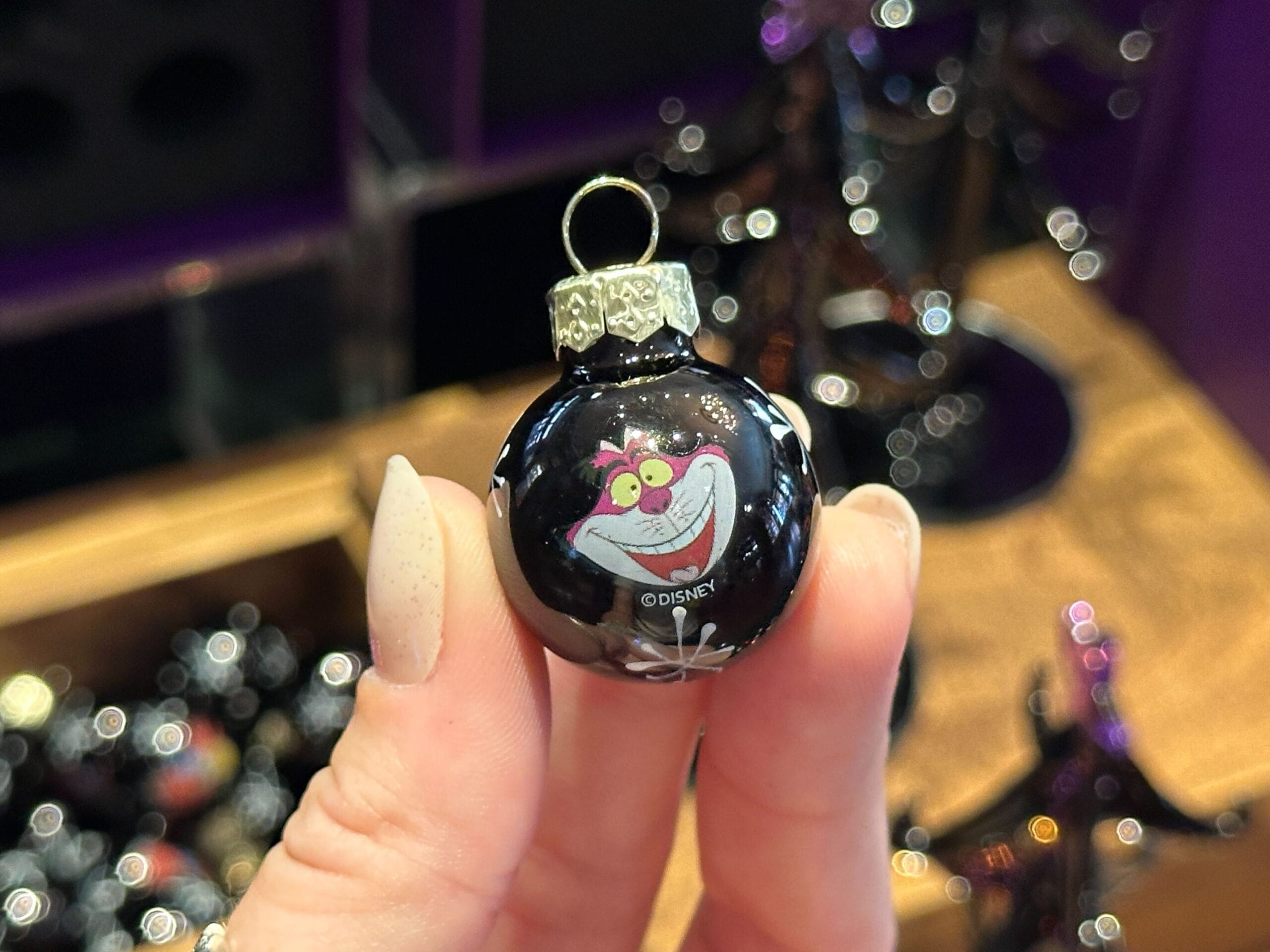 A hand holds a small black Villain Ornament featuring the Disney Cheshire Cat's smiling face.