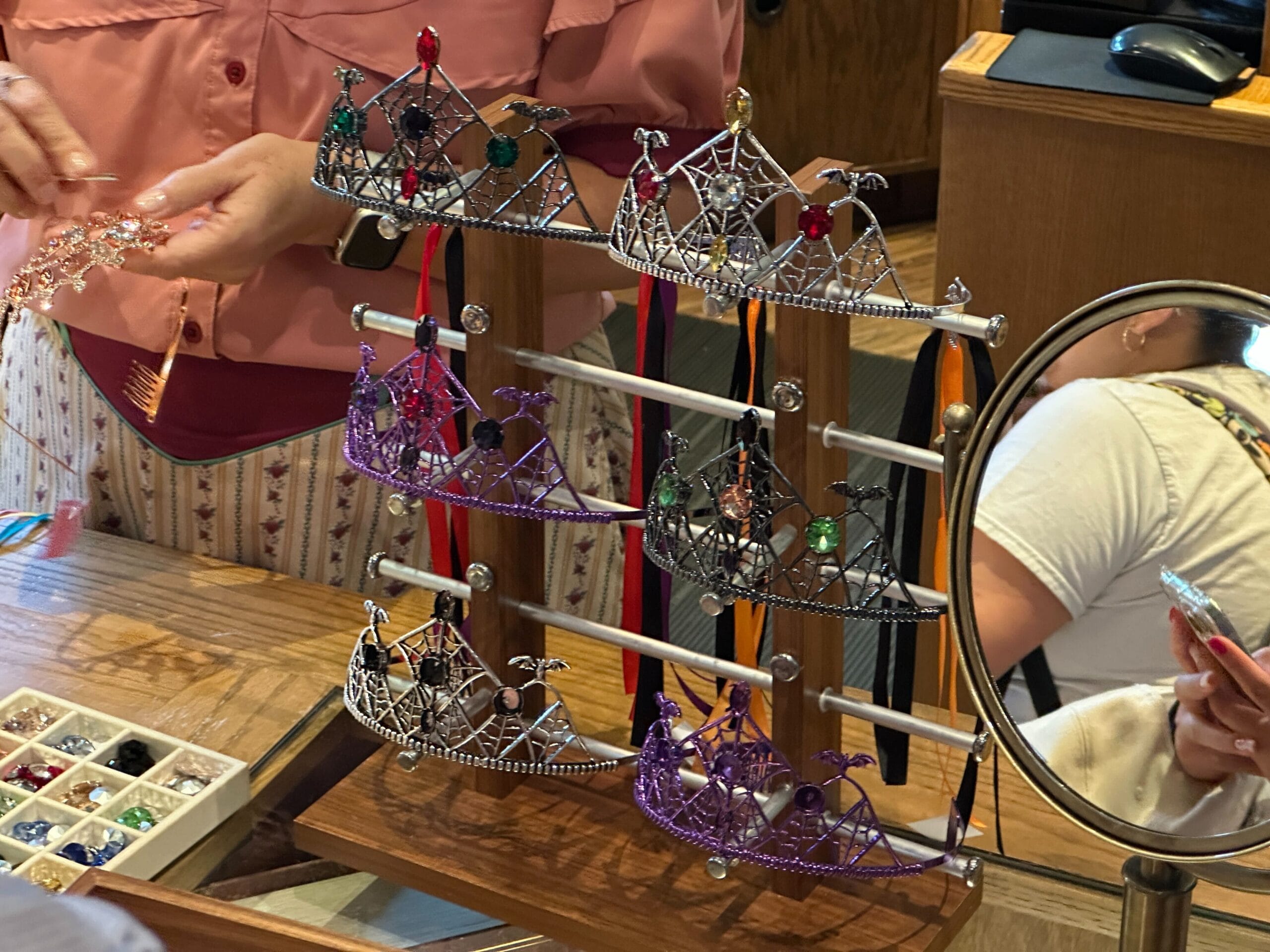 A person displays customizable jeweled tiaras on a stand, as another holds a phone near a mirror and gemstone box.