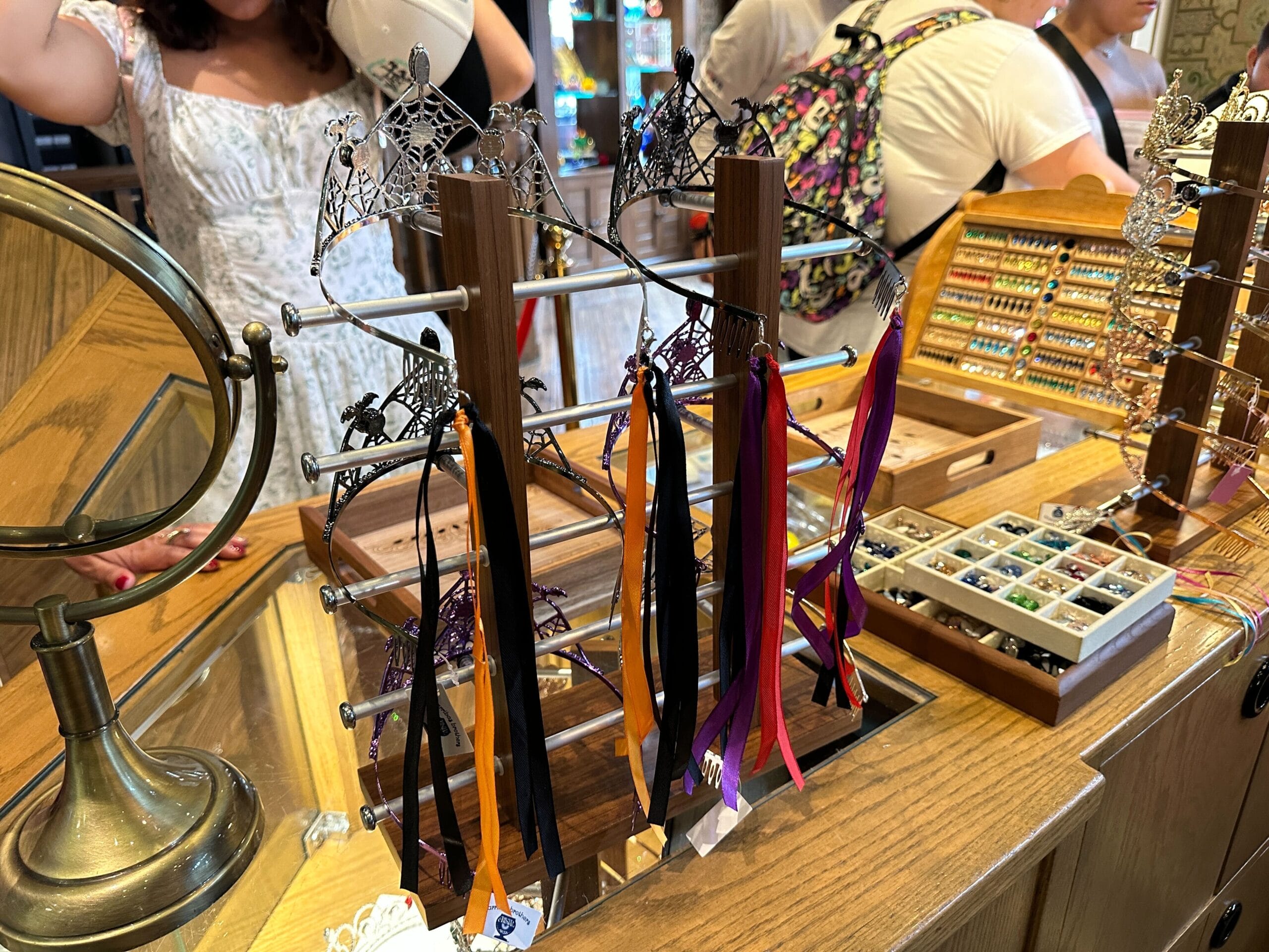 Display of customizable hair accessories with ribbons and Halloween tiaras on a wooden counter in a shop, people in background.