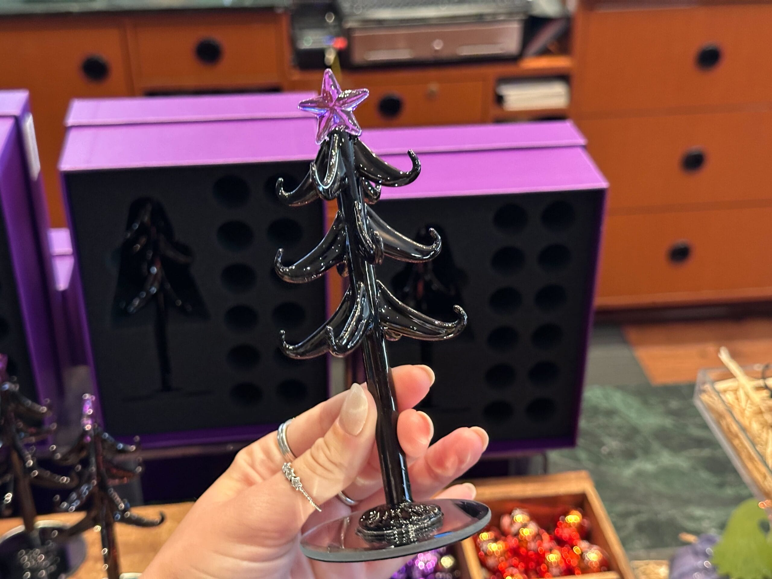 A hand holds a black Halloween Glass Tree with a purple star; villain ornaments and purple boxes in the background.