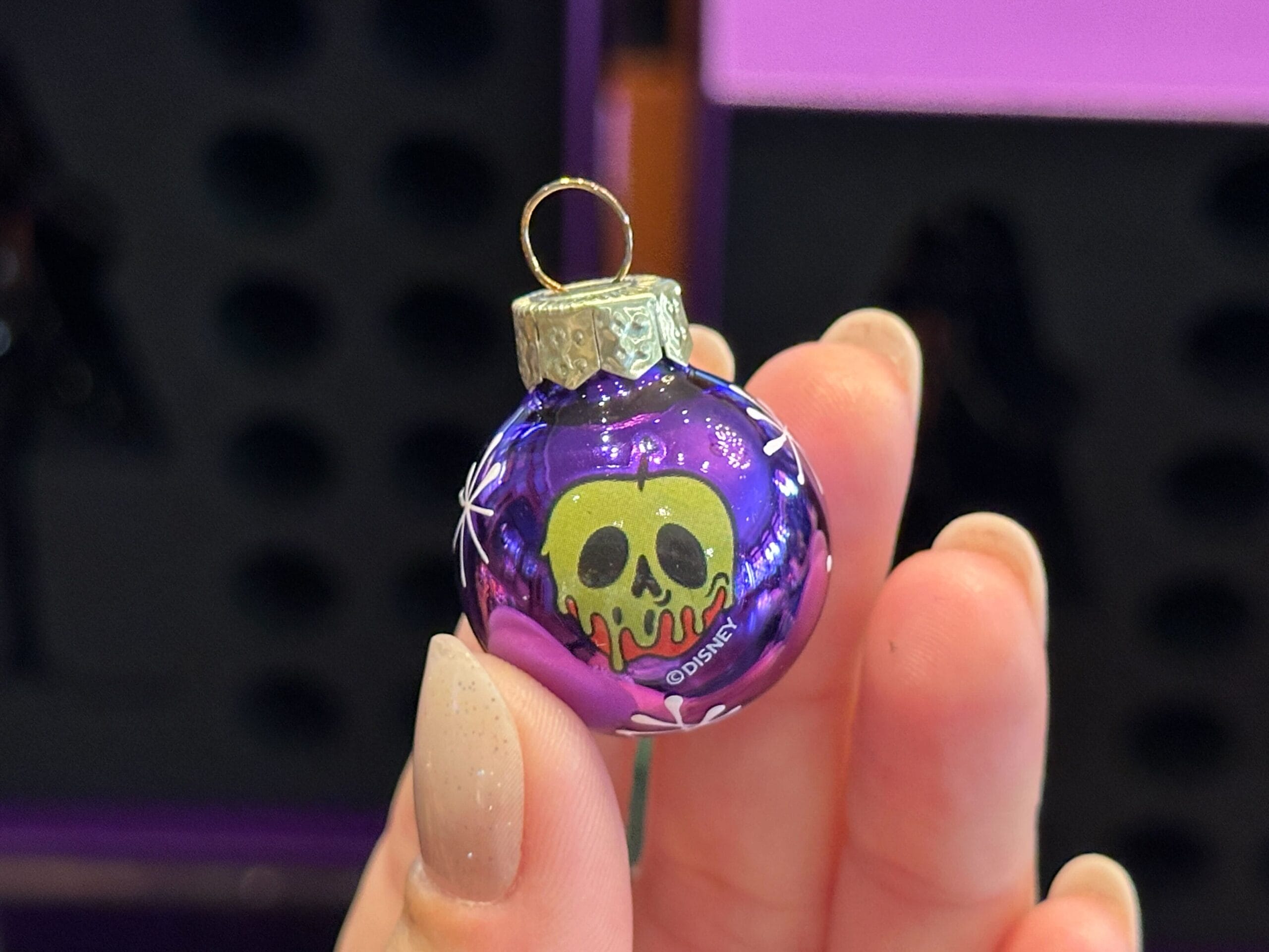 A hand holds a purple Villain Ornament featuring a green, dripping poison apple design from Disney’s Snow White.
