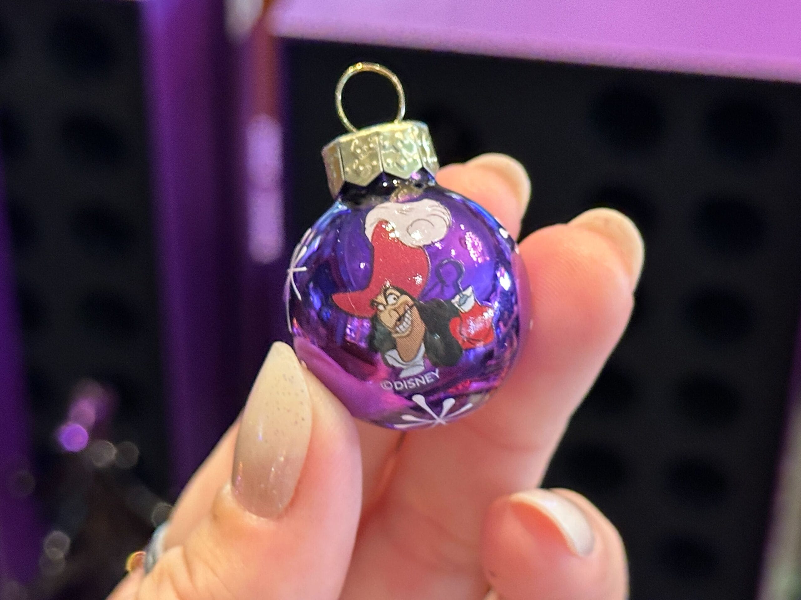 A hand holds a small purple Villain Ornament featuring Disney's Ursula, with a gold cap and snowflake details.