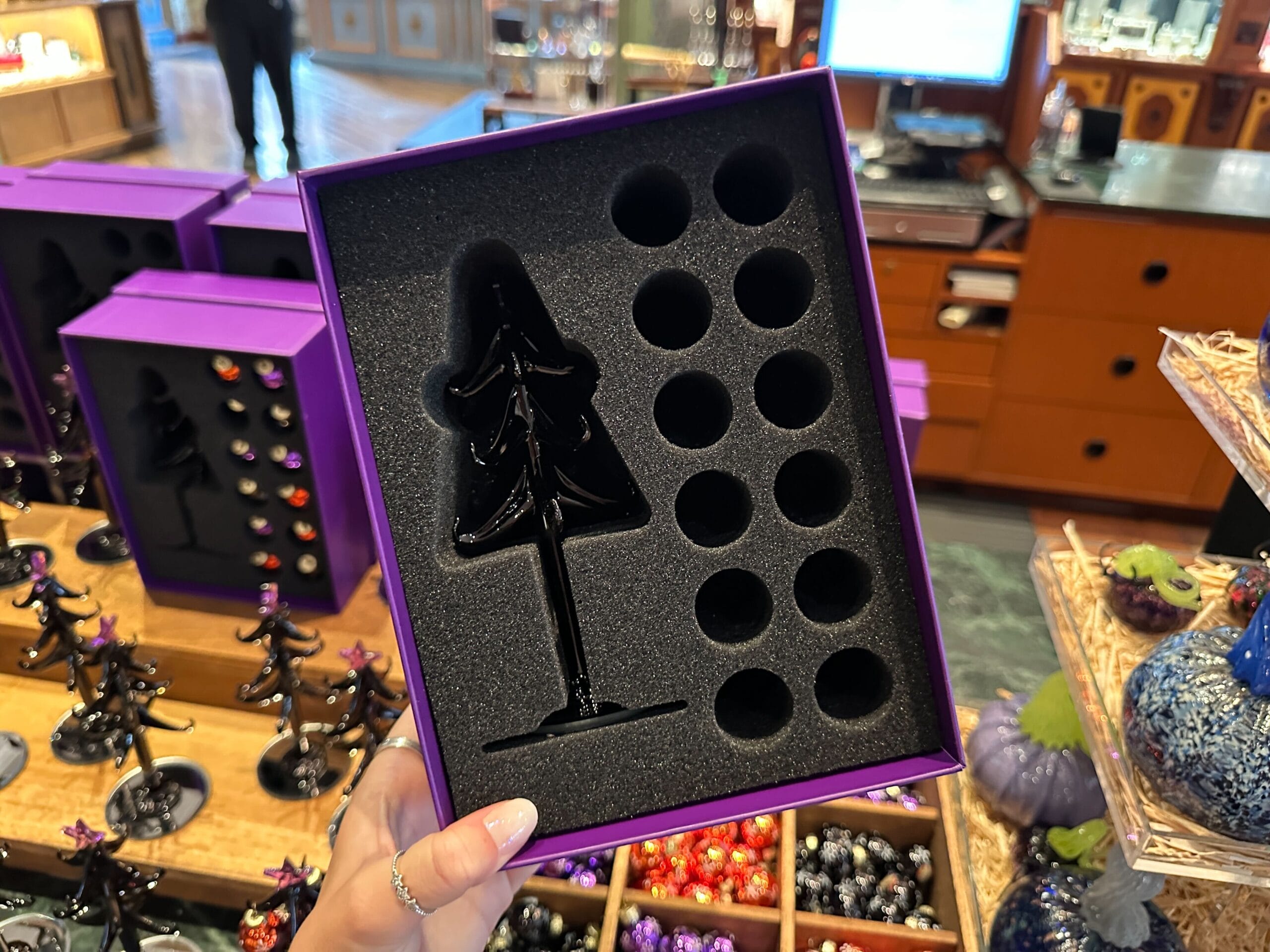 A hand holds an open box with a foam insert for Halloween Glass Trees and Villain Ornaments in a store display.
