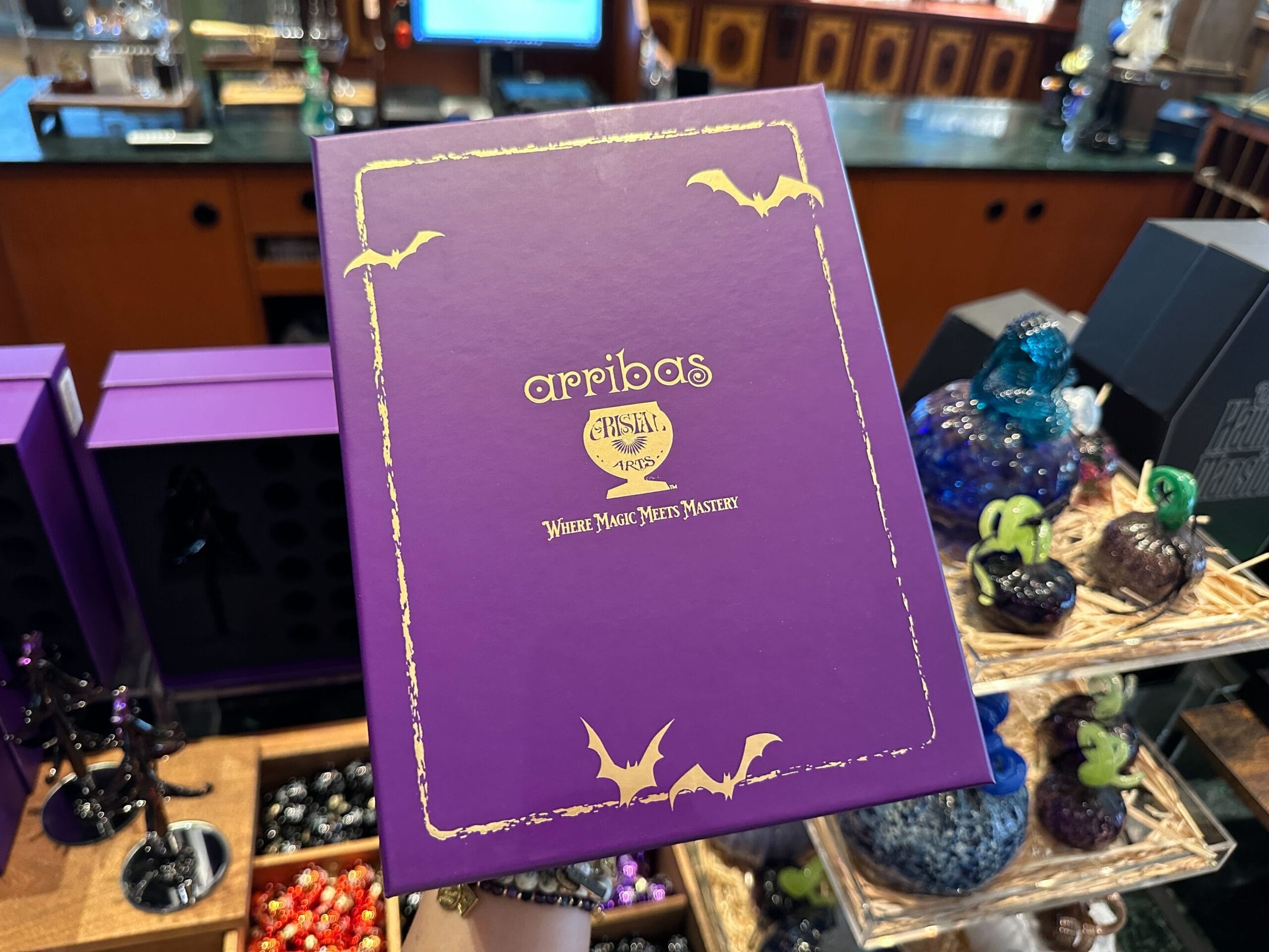 A purple Arribas gift box with gold bats, held in a Disney World store with Halloween Glass Trees in the background.