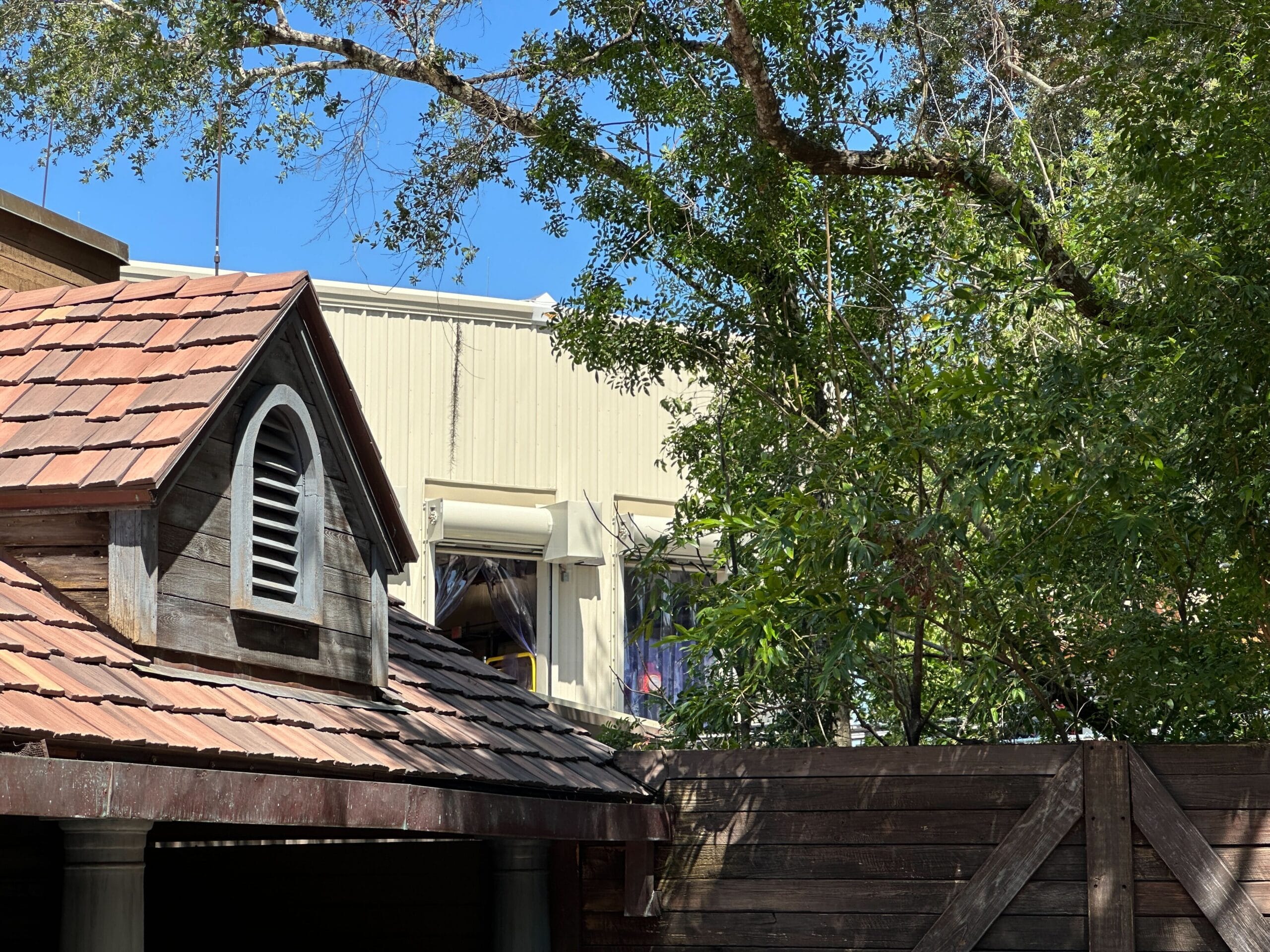 A wooden shed roof, high fence, and leafy trees hint at Big Thunder Mountain refurbishment under a clear blue sky.