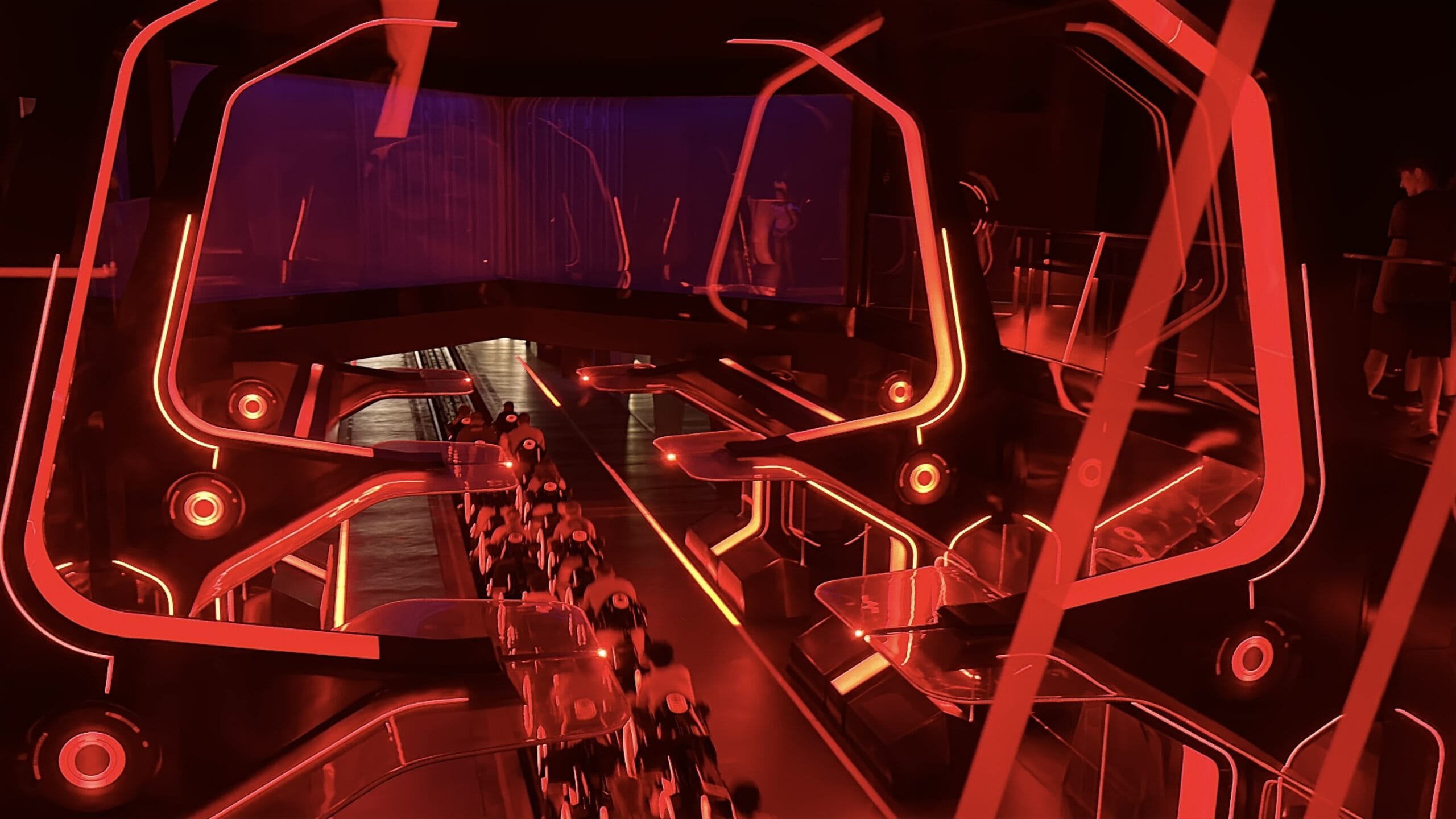 A futuristic, neon-lit indoor attraction with red lights and people riding TRON Lightcycle Run motorcycles on a glowing track.