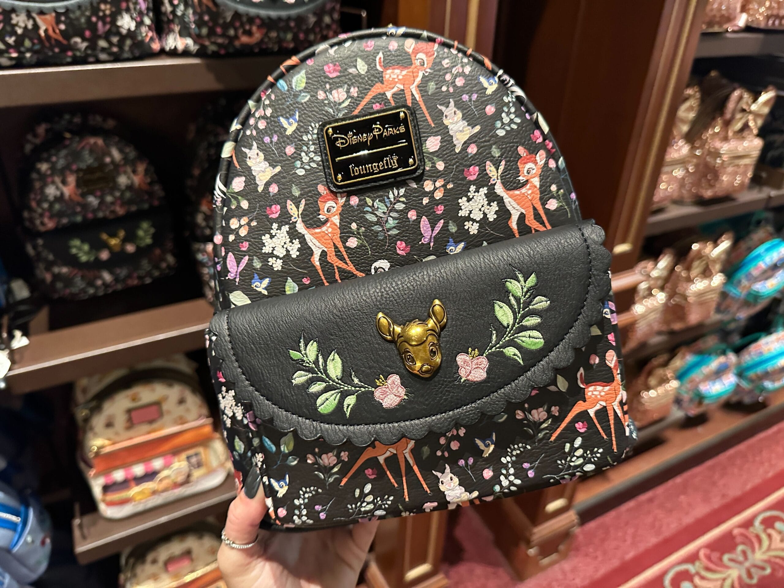 A hand holds a black Bambi Loungefly mini backpack with floral patterns on a Disney World store display.