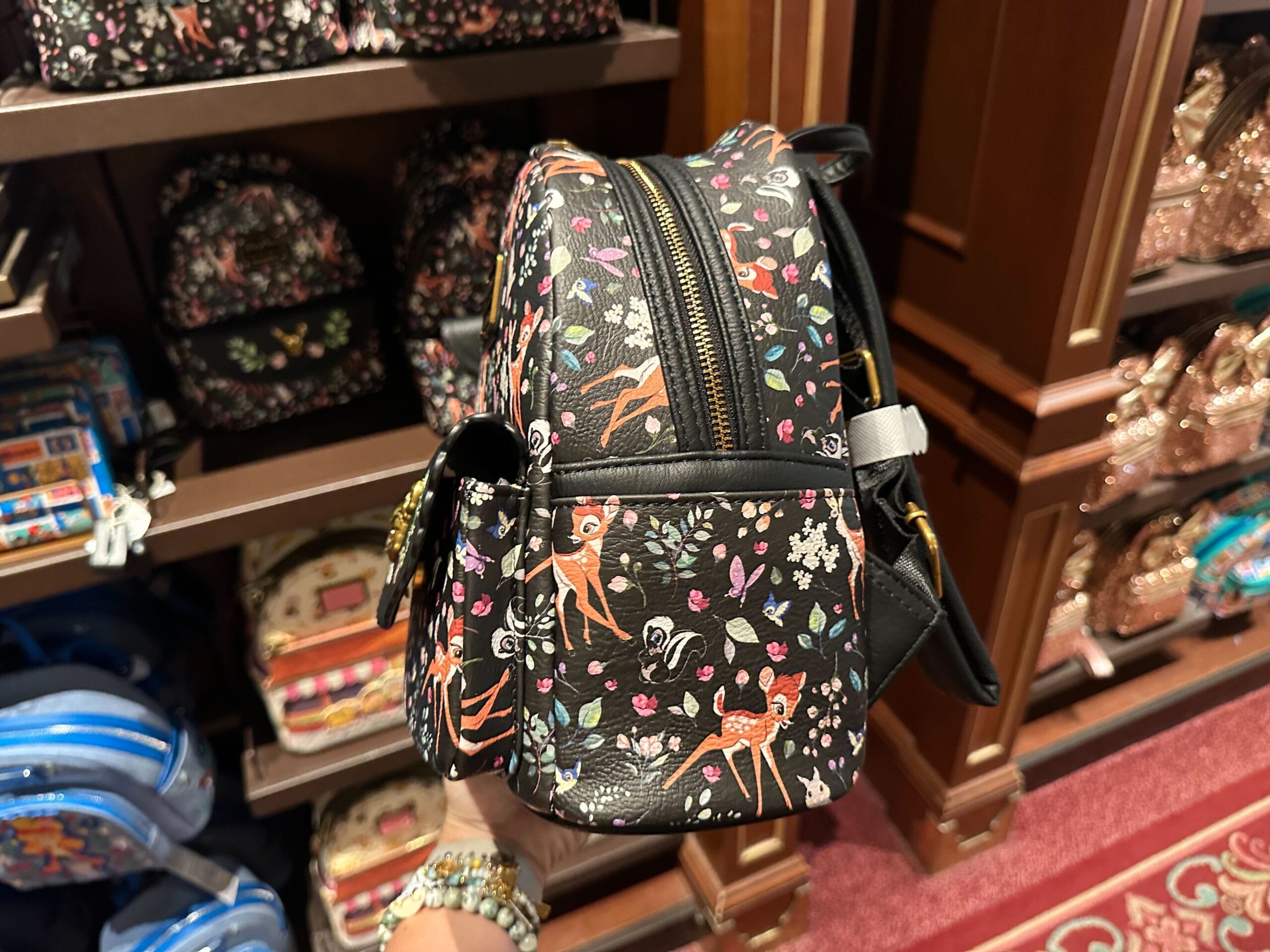 A hand holds a Bambi Loungefly Mini Backpack with floral designs at a Disney World store display of patterned bags.