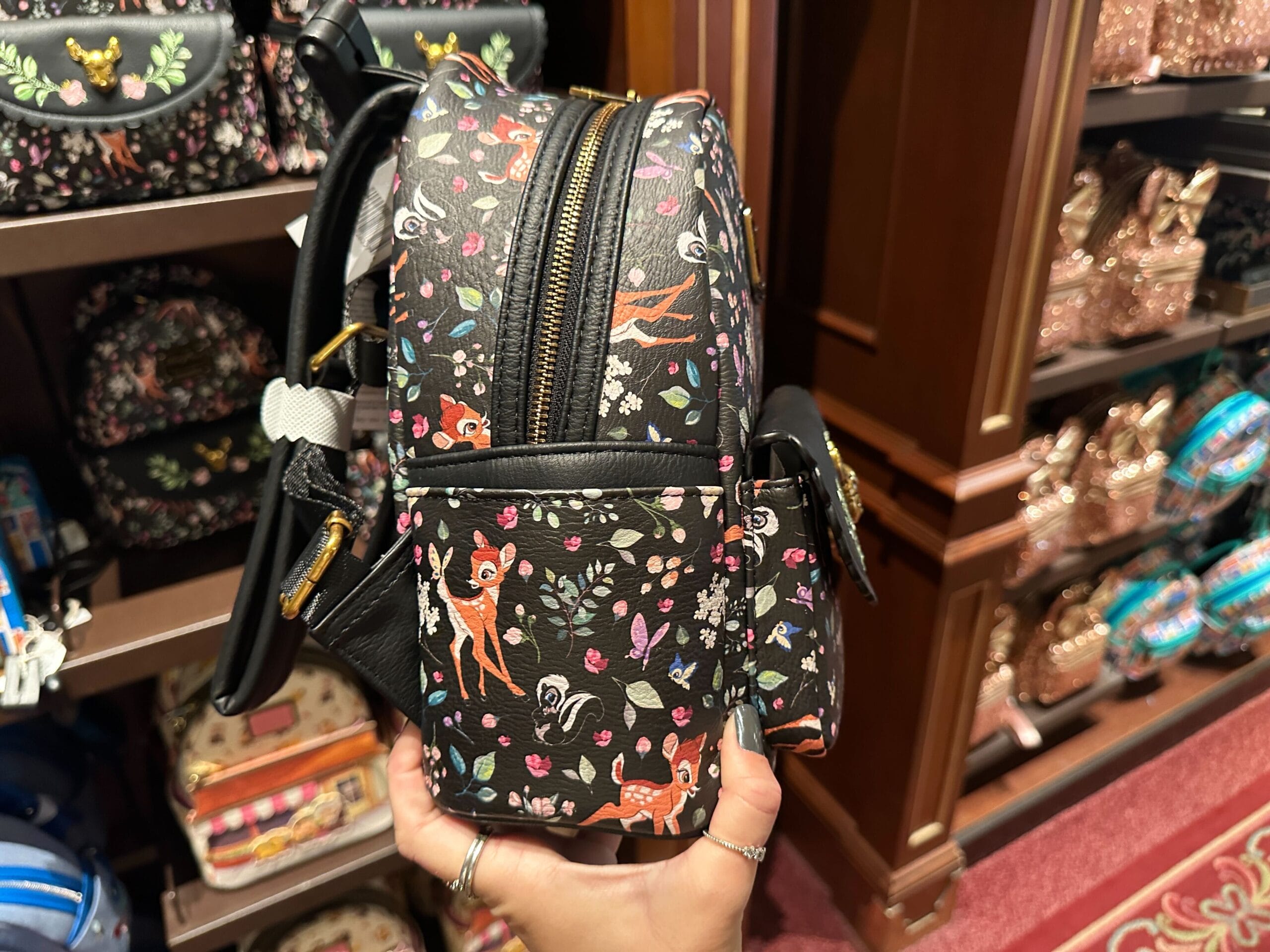 A hand holds a Bambi Loungefly mini backpack with floral prints, displayed among other patterned bags in a Disney World store.