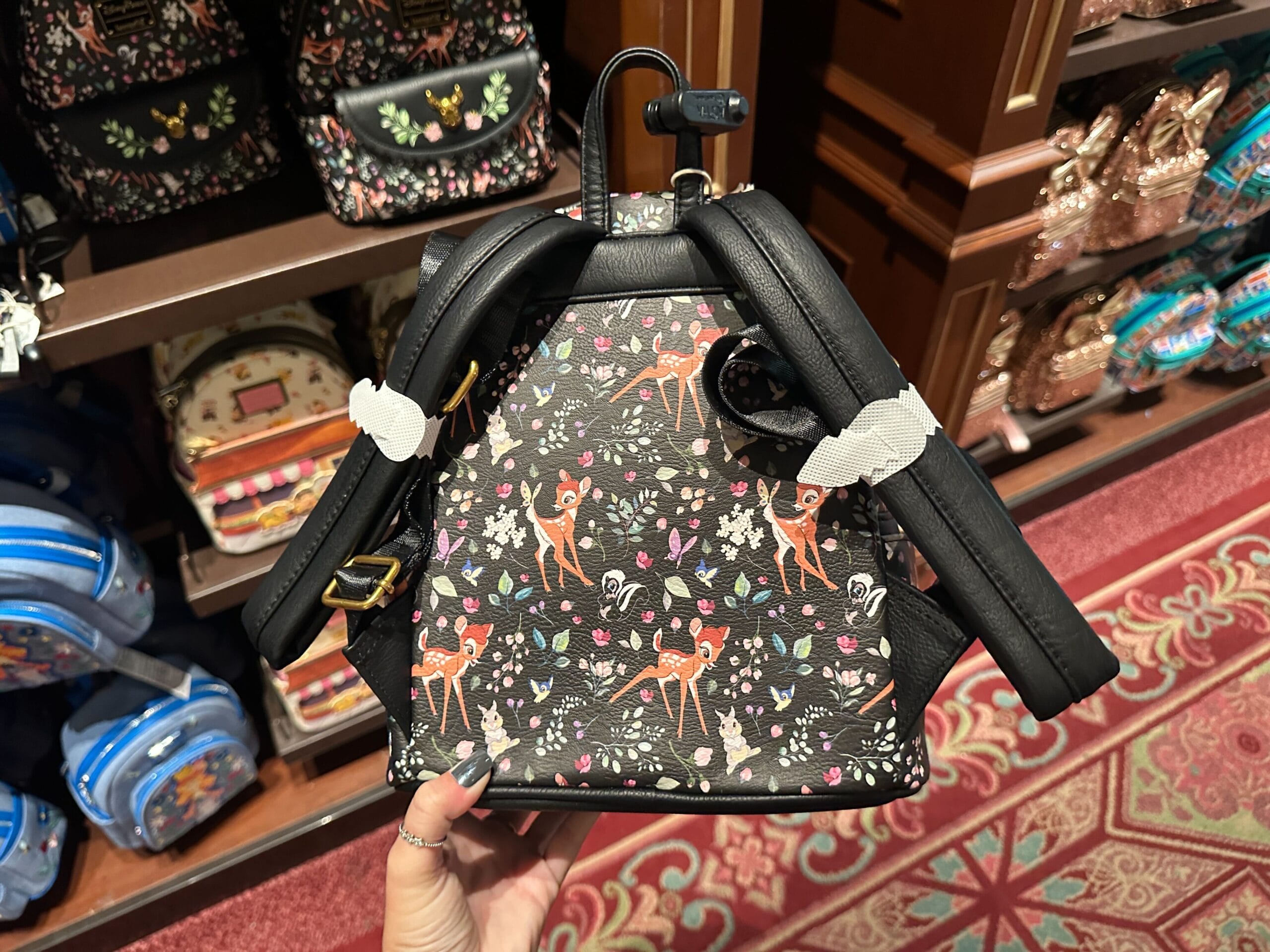 A hand holds a Bambi Loungefly mini backpack with floral designs in a store filled with colorful Disney World backpacks.