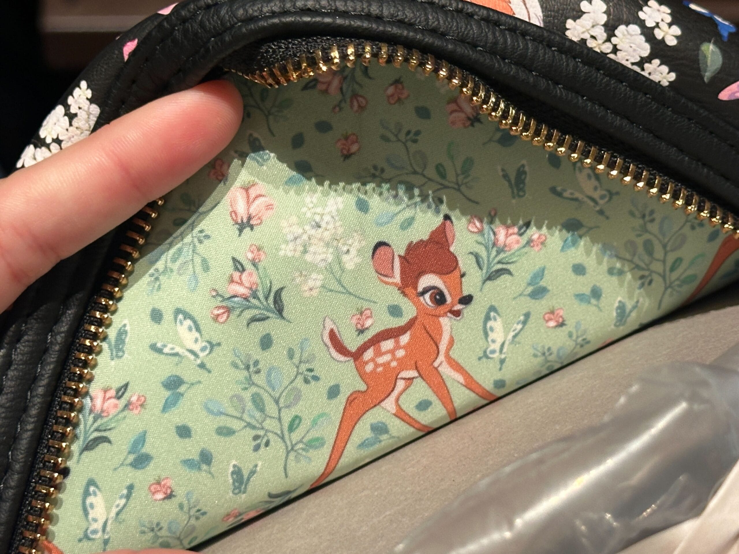 A hand opens a purse to reveal a Bambi Loungefly Mini Backpack lining with cartoon fawn, butterflies, and greenery.