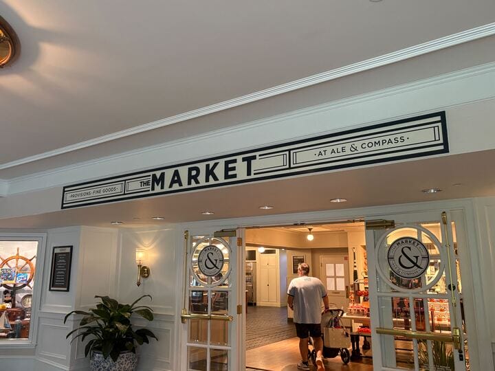Entrance to "The Market at Ale & Compass" with glass doors and a man pushing a cart inside a well-lit shop.