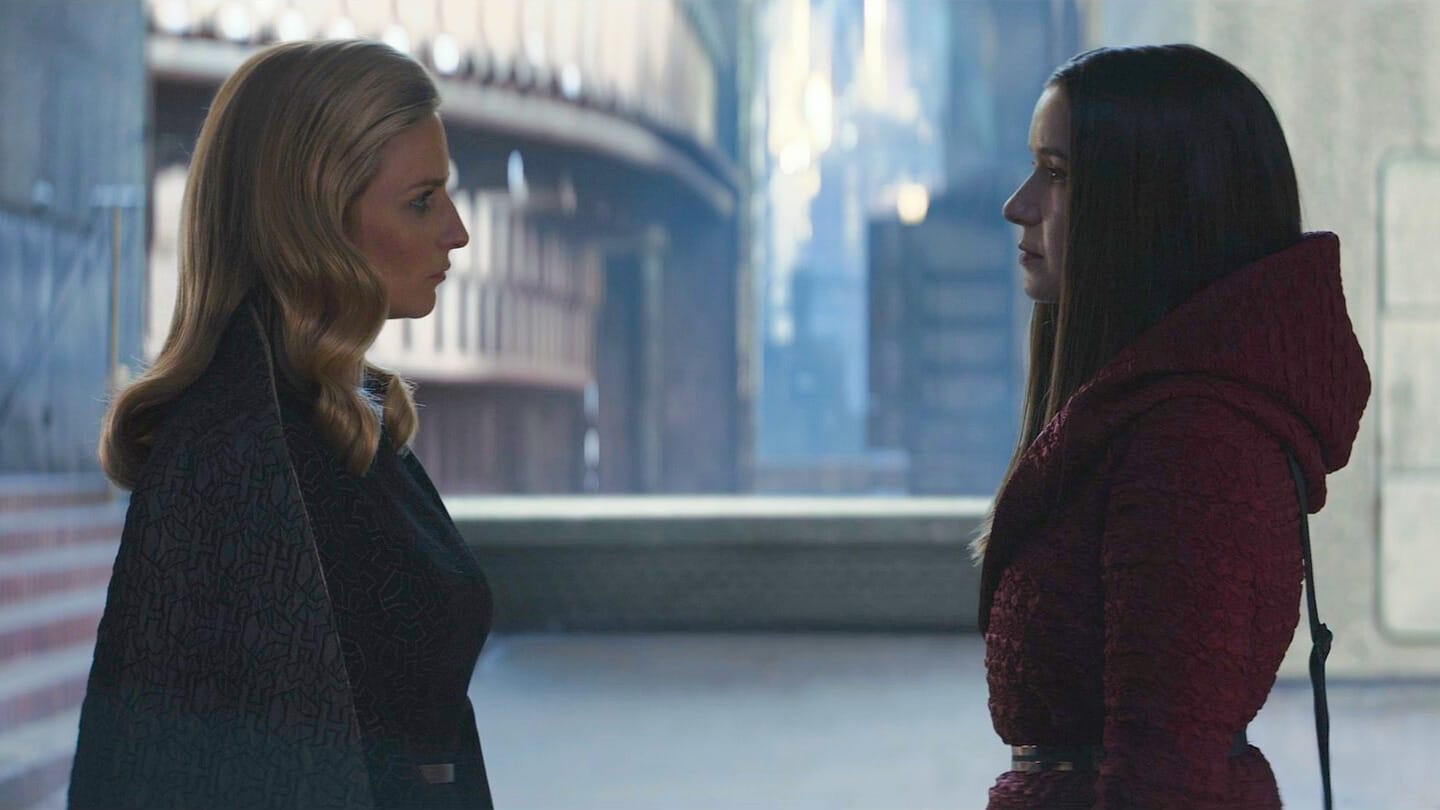 Two women face each other in a dim, futuristic hallway, dressed in dark formals—a scene reminiscent of Andor's style.