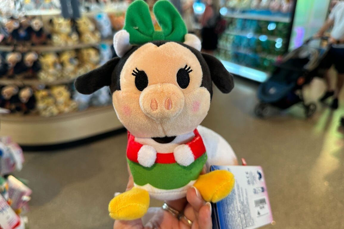 Plush toy resembling Clarabelle cow with a green bow, held in a store with shelves of plushies at Walt Disney World.