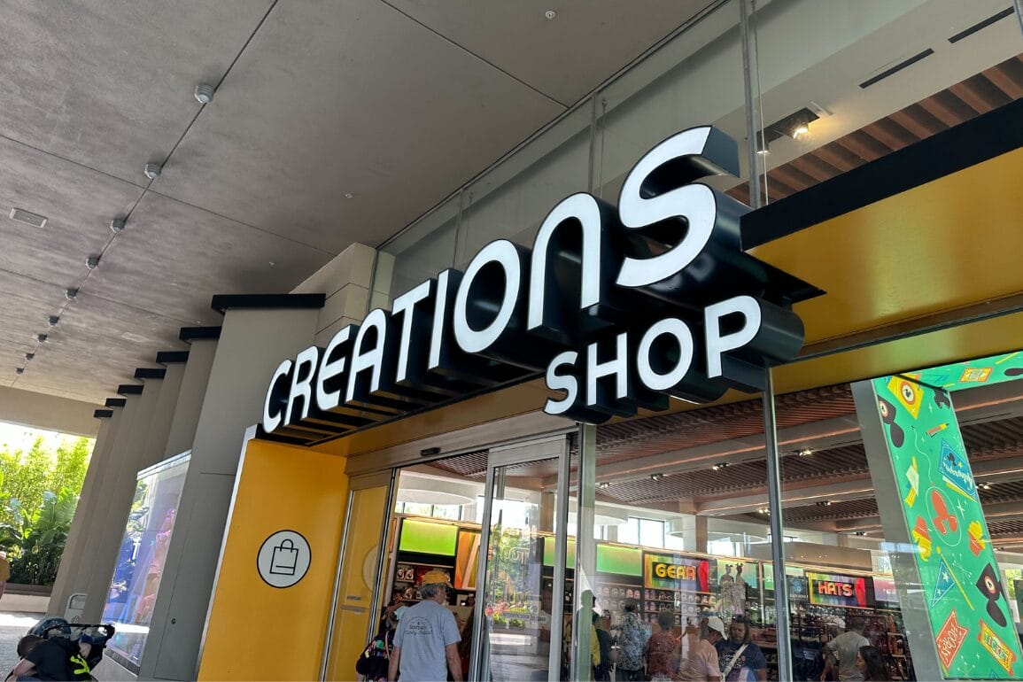 Entrance of Creations Shop at Walt Disney World, with a large sign and shopping bag icon above glass doors.