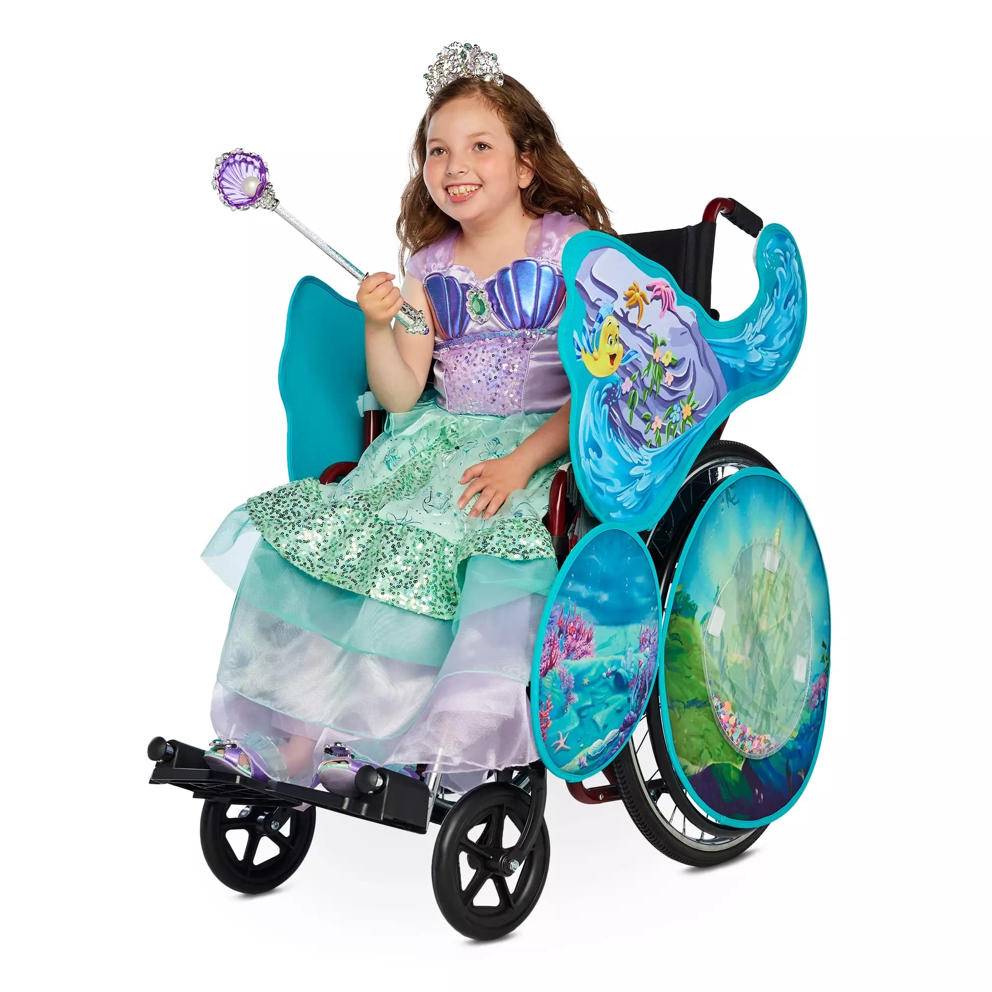Smiling girl in a mermaid costume from Disney Store holds a wand and sits in a wheelchair with ocean-themed panels.