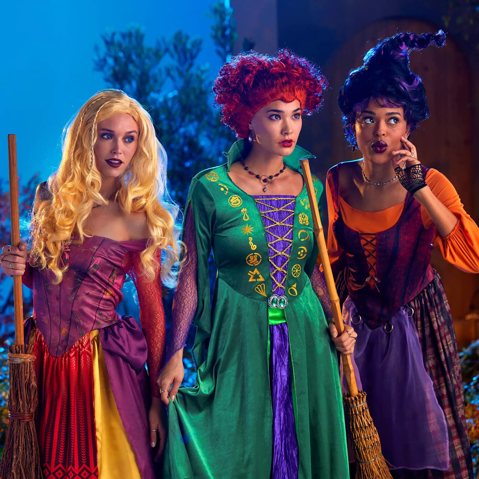 Three women in colorful Disney Store Halloween costumes hold brooms, standing together and looking surprised or curious.