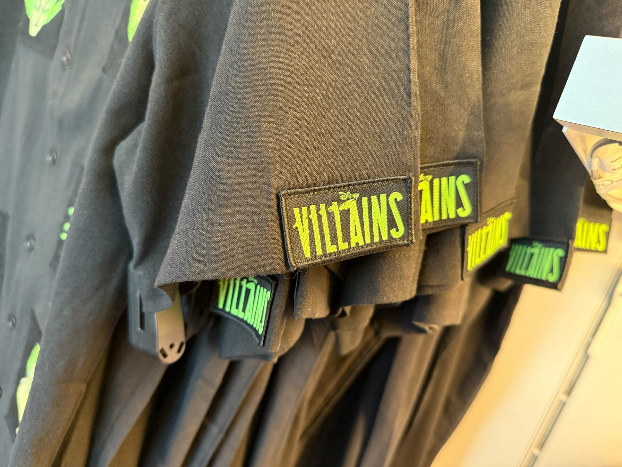 Black shirts with green "Villains" patches hanging on a rack—perfect Halloween merchandise at Disneyland.