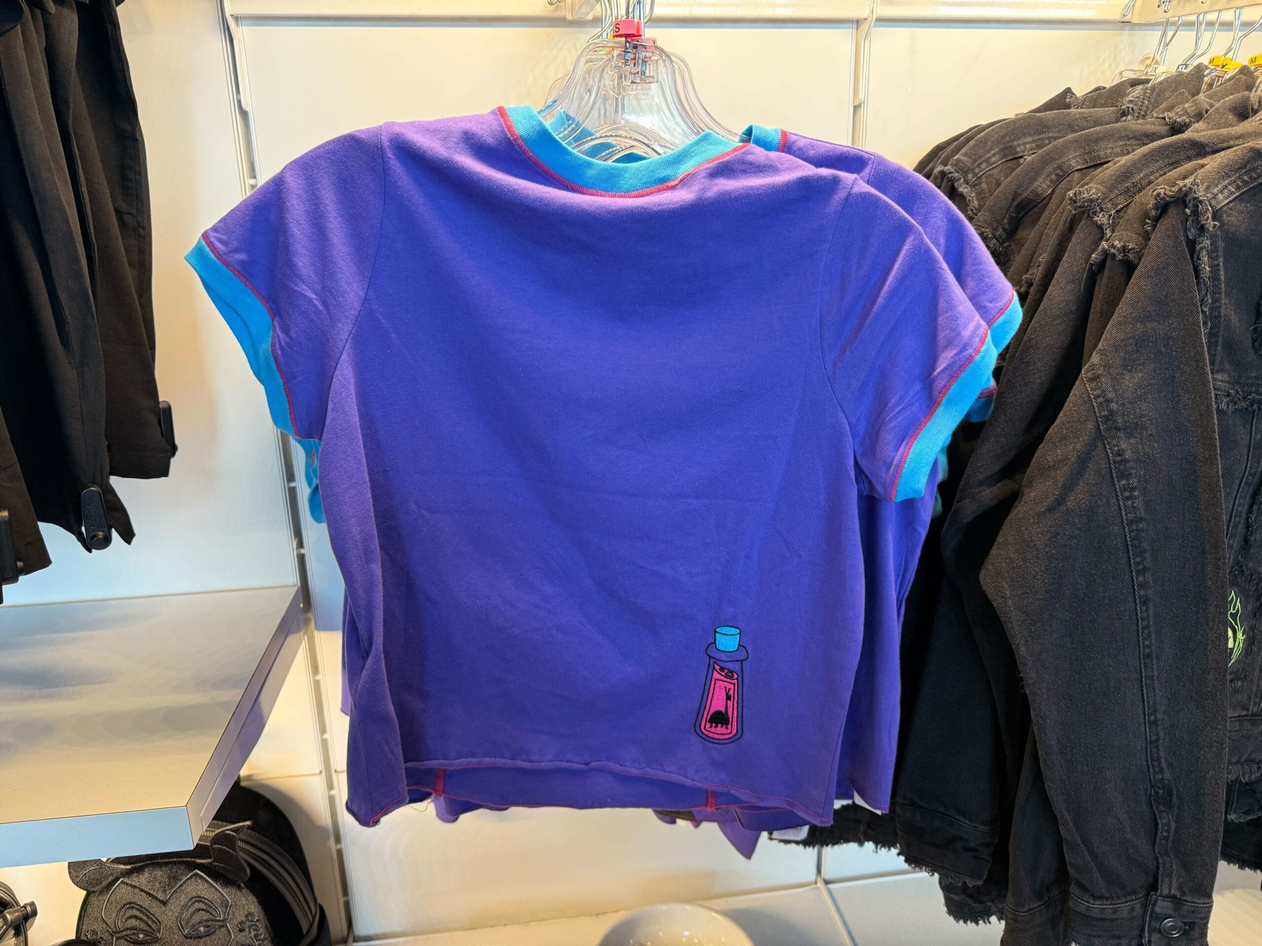Purple t-shirt with blue trim, featuring a small bottle graphic, hangs near black jeans—perfect for Villains merchandise lovers.