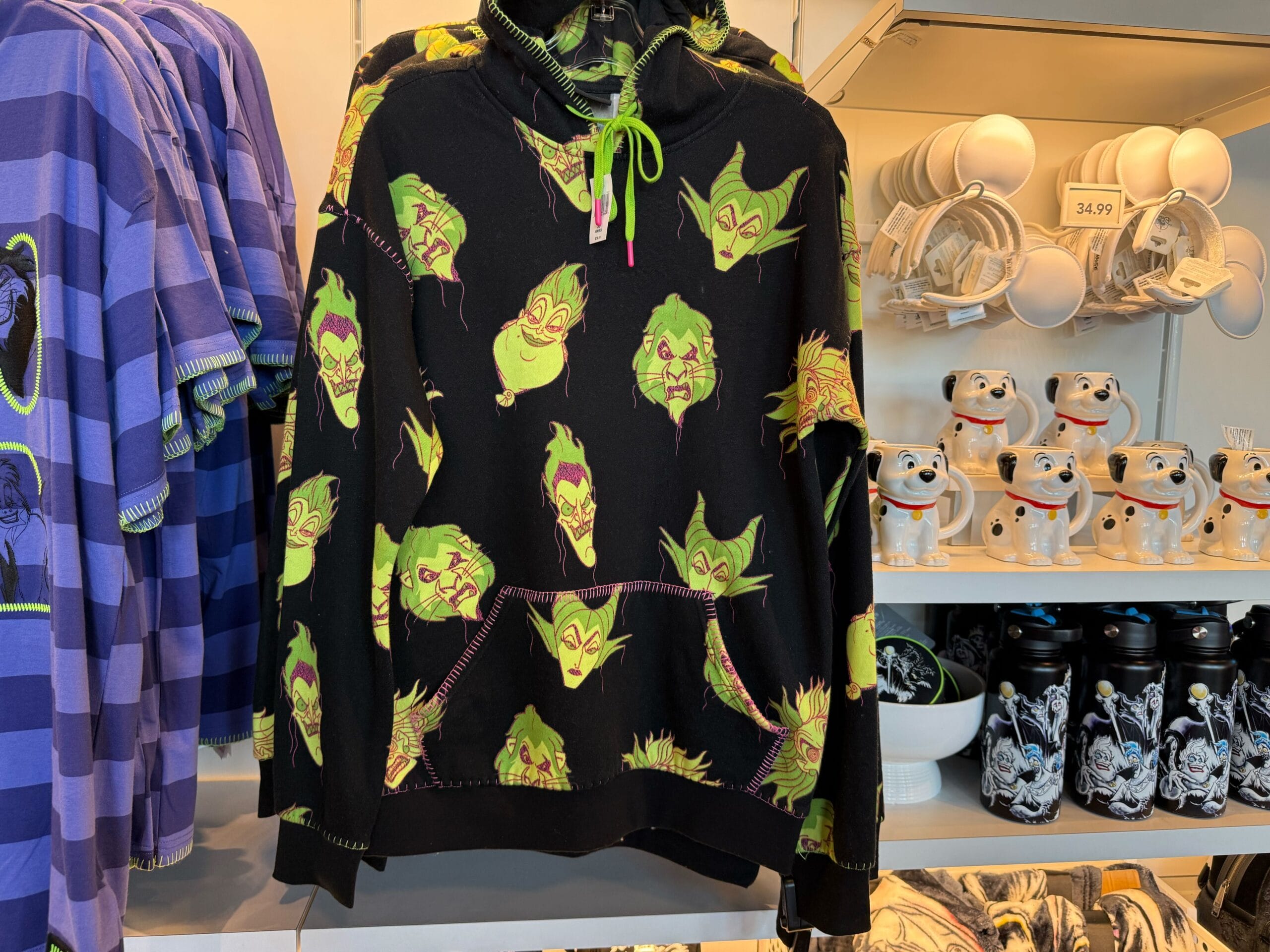 Black hoodie with green evil face patterns hangs in a store, surrounded by Villains Merchandise shirts, mugs, and water bottles.