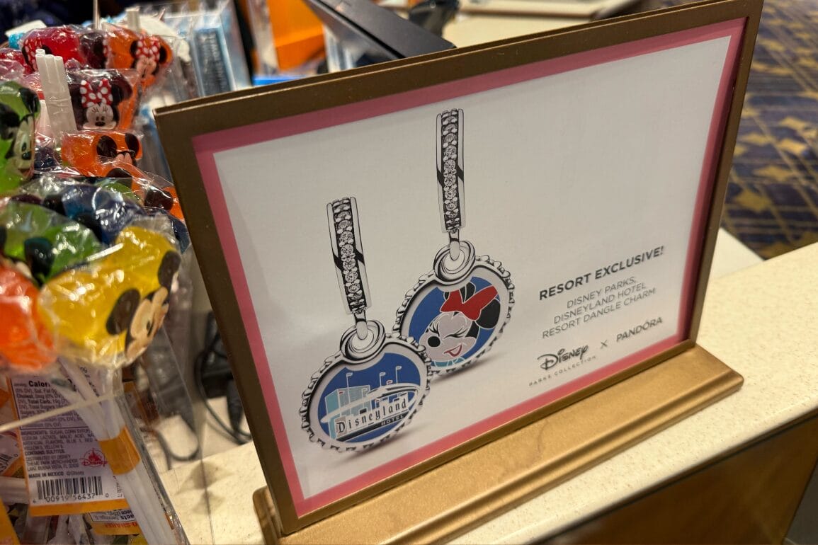 Sign advertising exclusive Disneyland Resort Pandora Charms next to colorful Mickey Mouse lollipops on the counter.