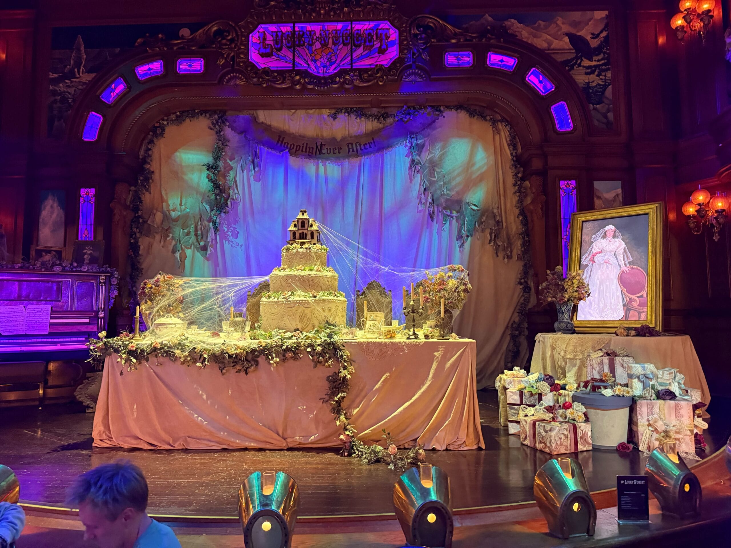 A stage at Phantom Manor with a grand cake, cobwebs, wedding gifts, and a bride's portrait in moody lighting.