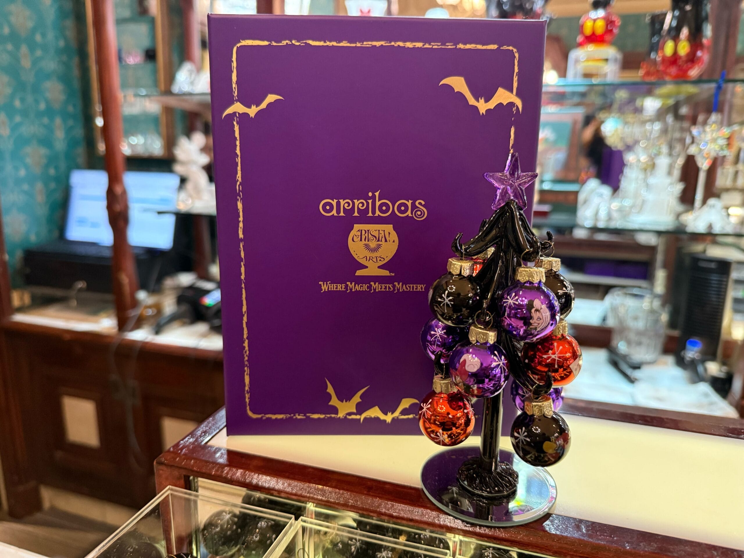 A small black Custom Glass Halloween Villains Tree with purple and orange ornaments stands in front of a purple Arribas Paris box.