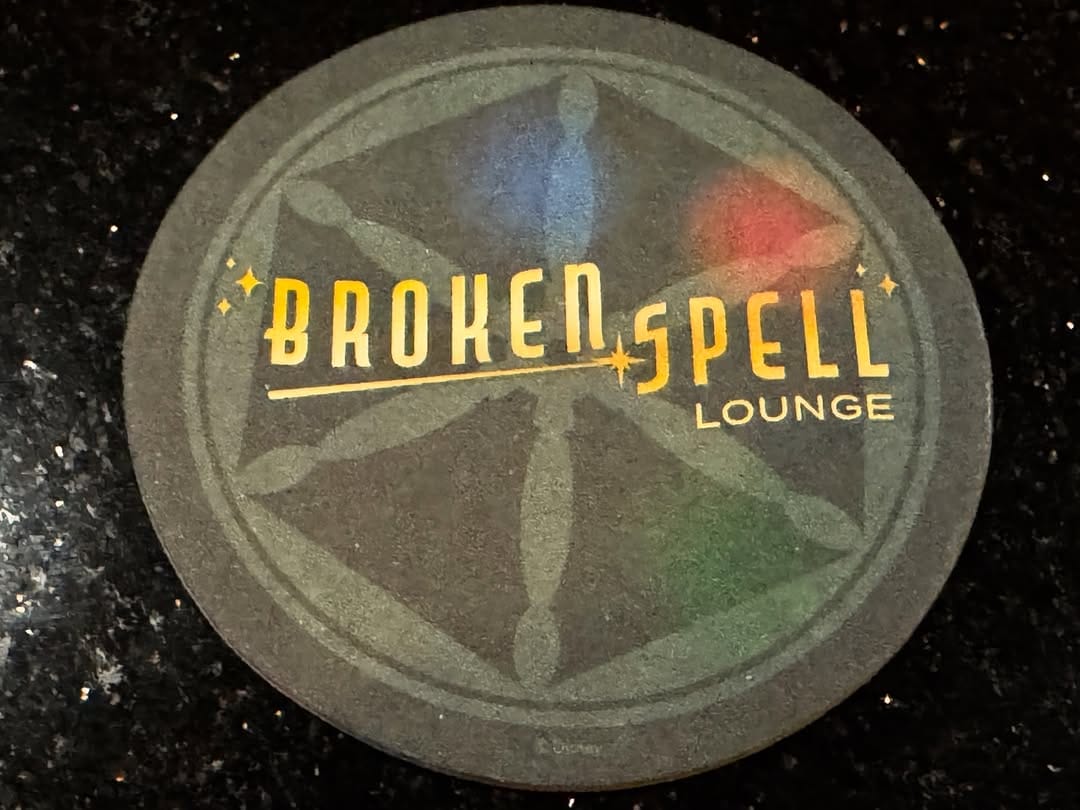 A round coaster with "Broken Spell Lounge" text and a mystical symbol on a dark background.