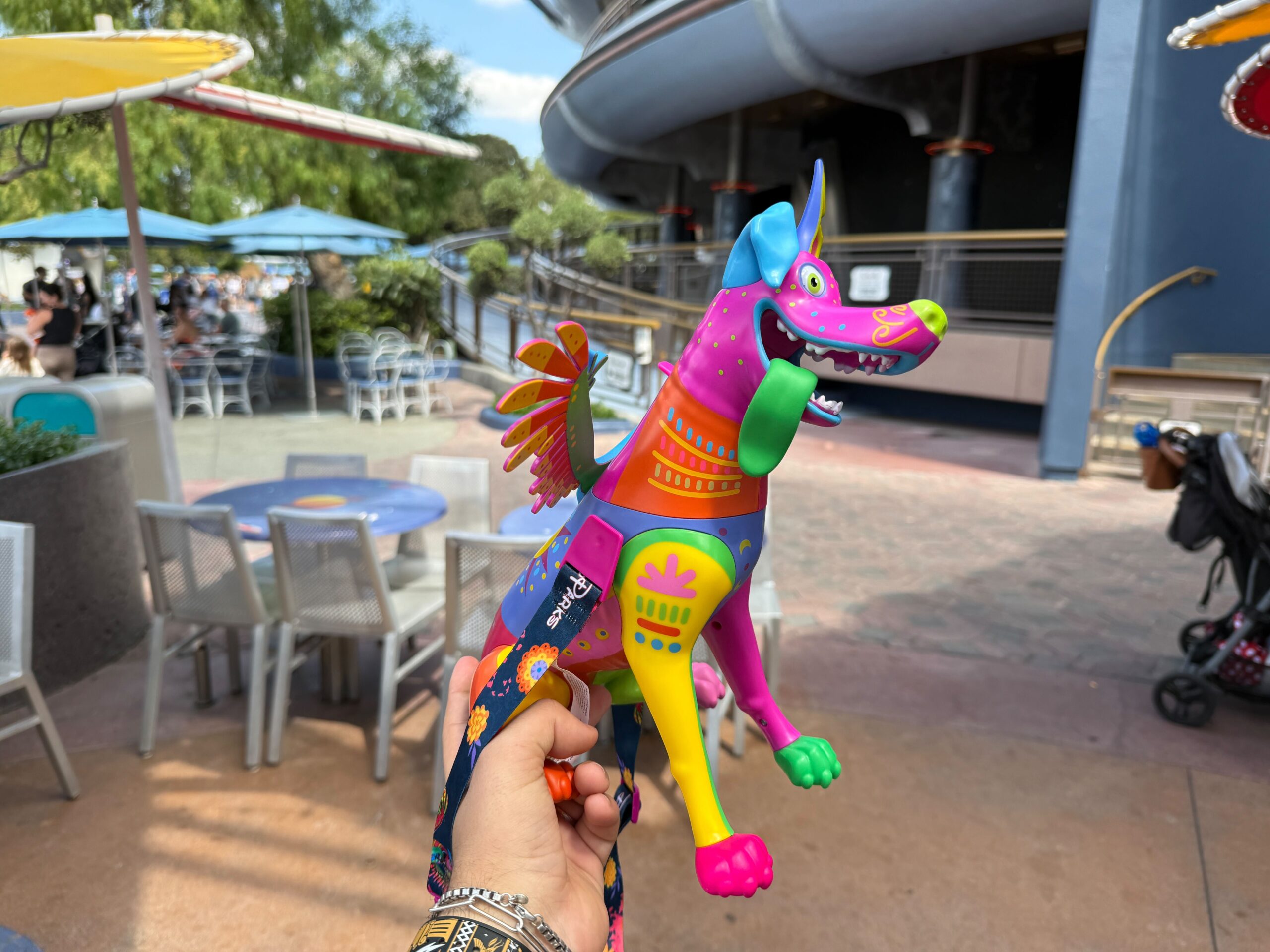 A hand holds a colorful Dante Sipper dog toy outside at a cafe with blue tables and umbrellas.