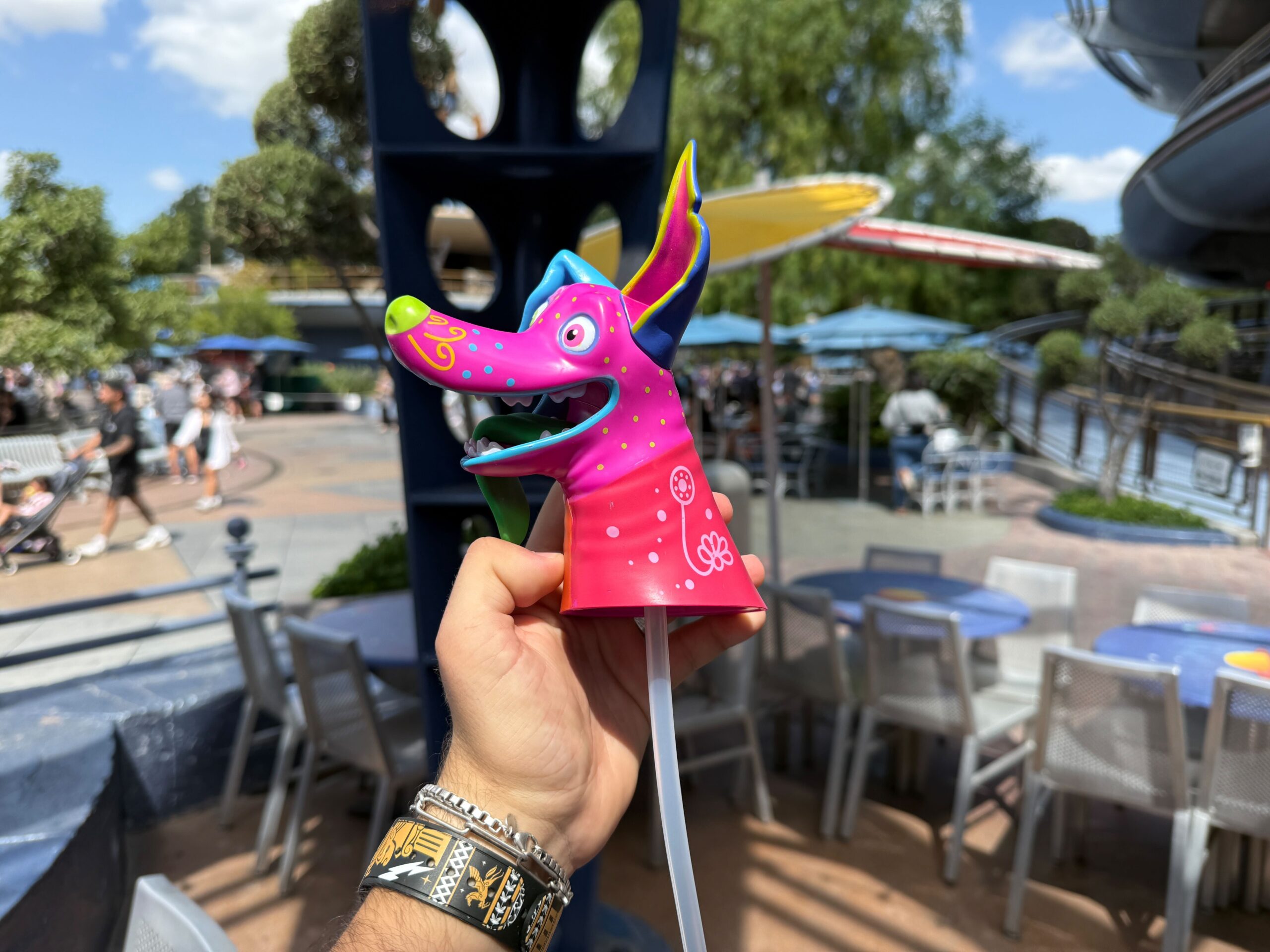 A colorful Dante Sipper dog-shaped cup is held up outdoors at Disneyland Resort, with tables and people in the background.