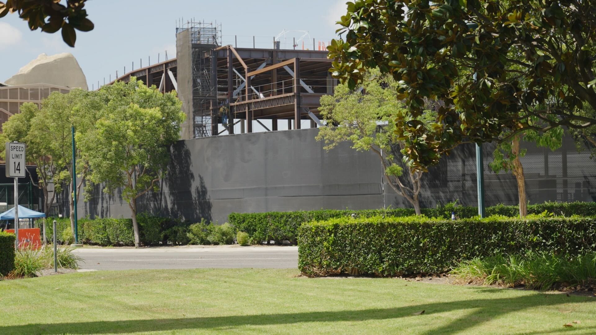 Steel structure under construction behind trees and hedges, with a speed limit sign and clear blue sky.