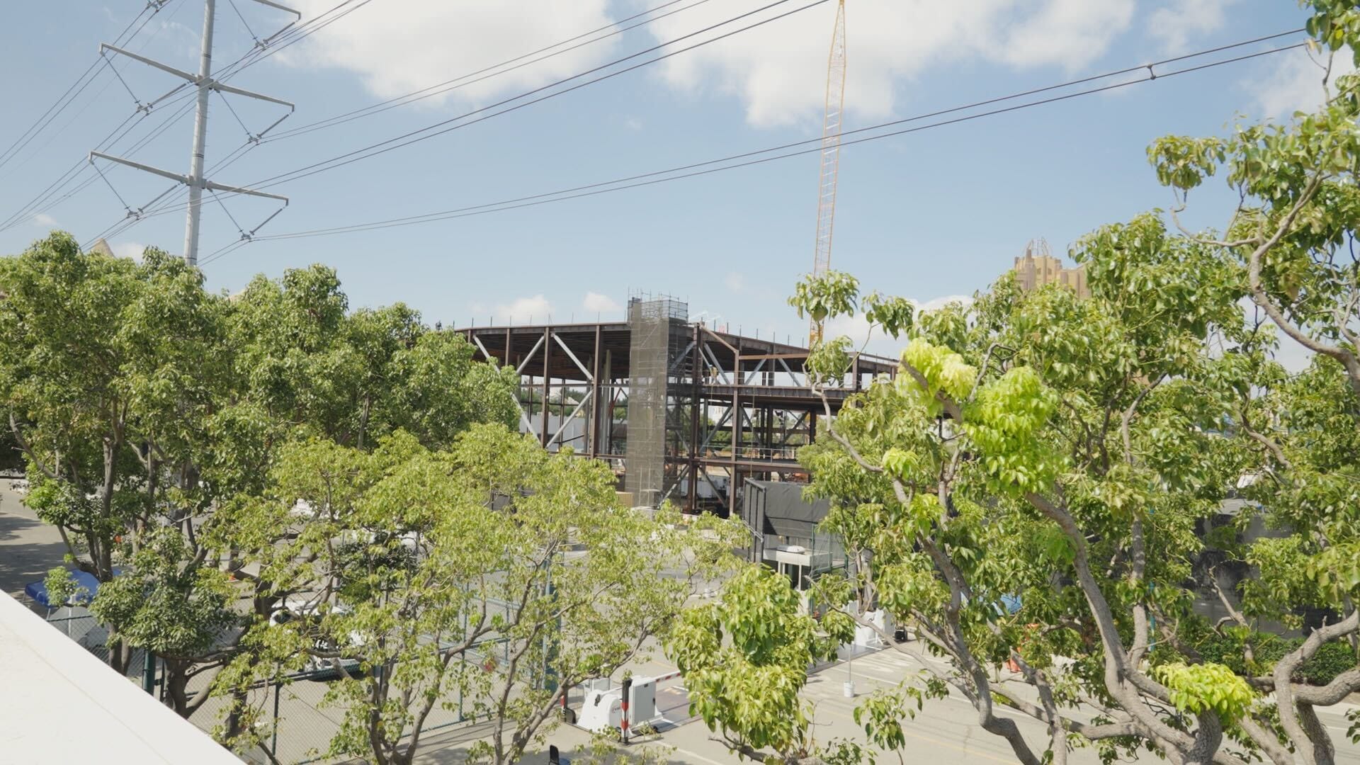 Steel structure under construction behind green trees, with power lines and a crane visible under a partly cloudy sky.