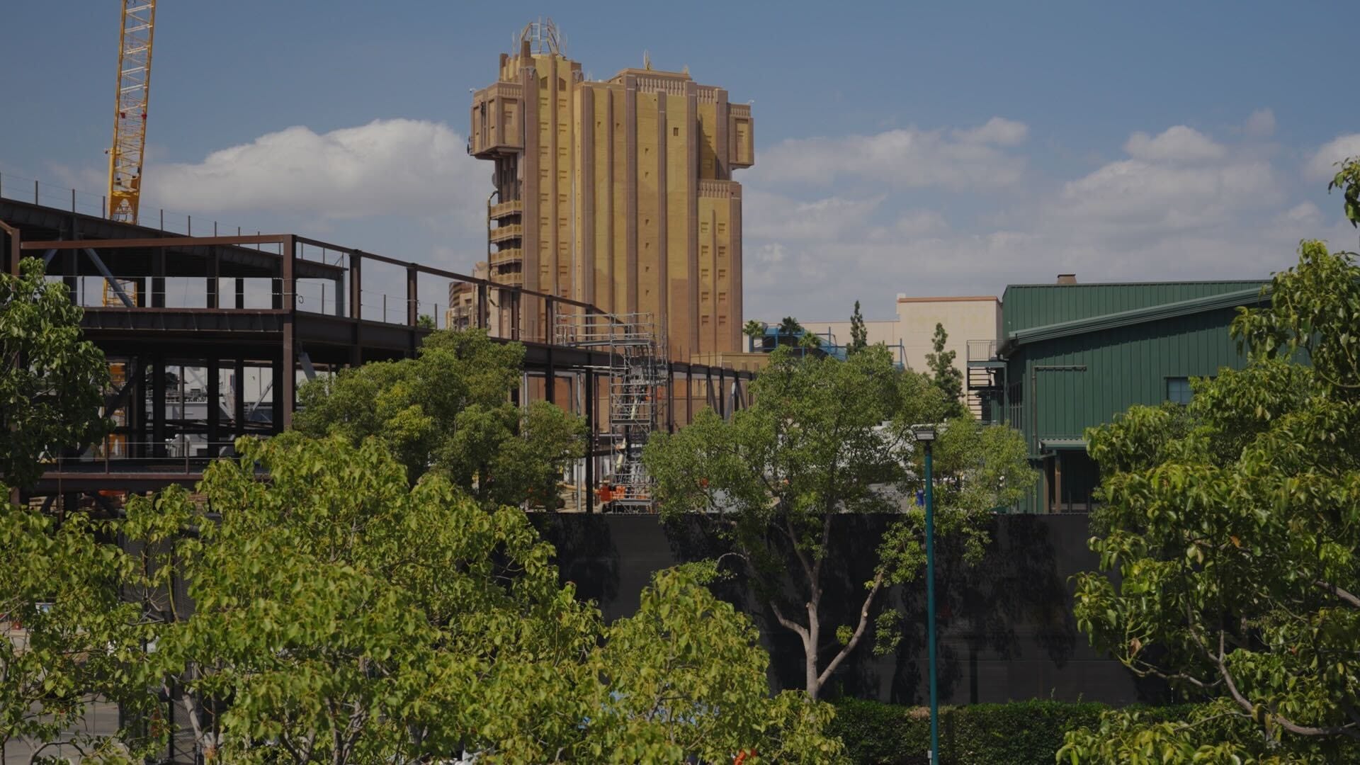 Brown tower rises behind trees and construction structures under a partly cloudy sky.