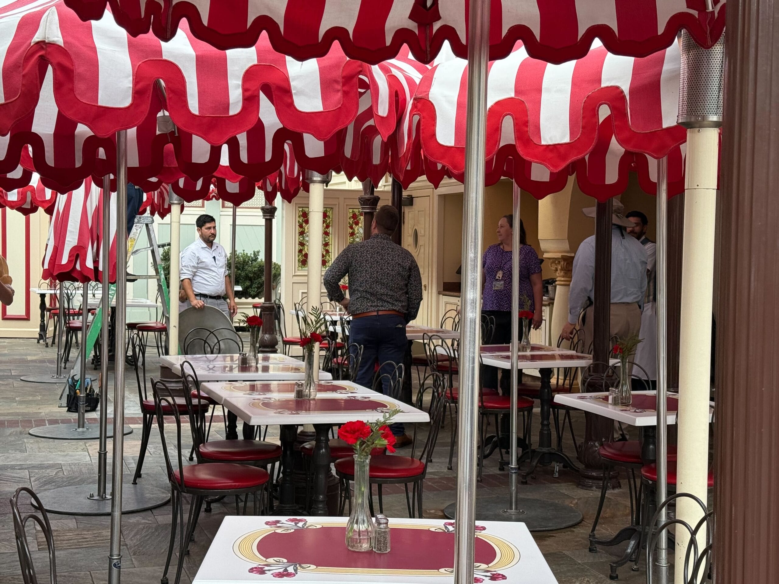 Outdoor cafe with red and white umbrellas, empty tables, and several people standing and talking in the background.