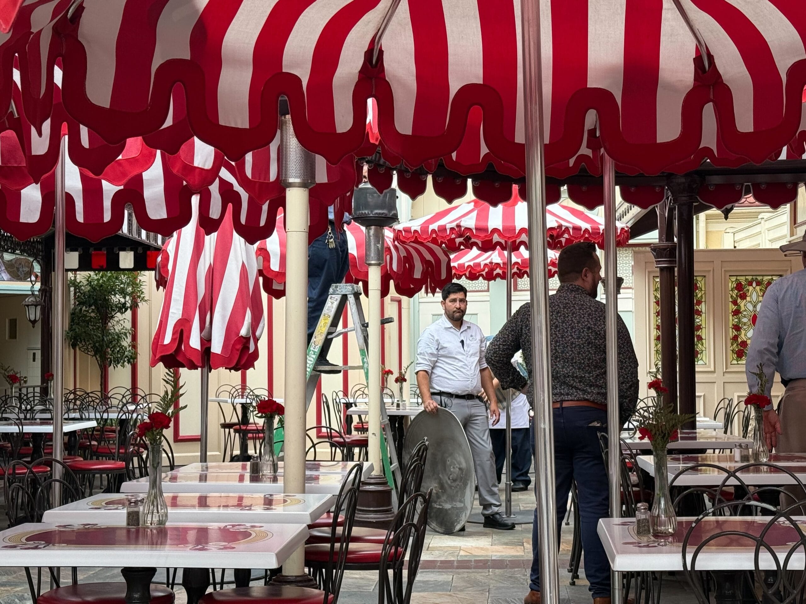 Several men set up under red and white striped umbrellas at an outdoor cafe with empty tables and chairs.