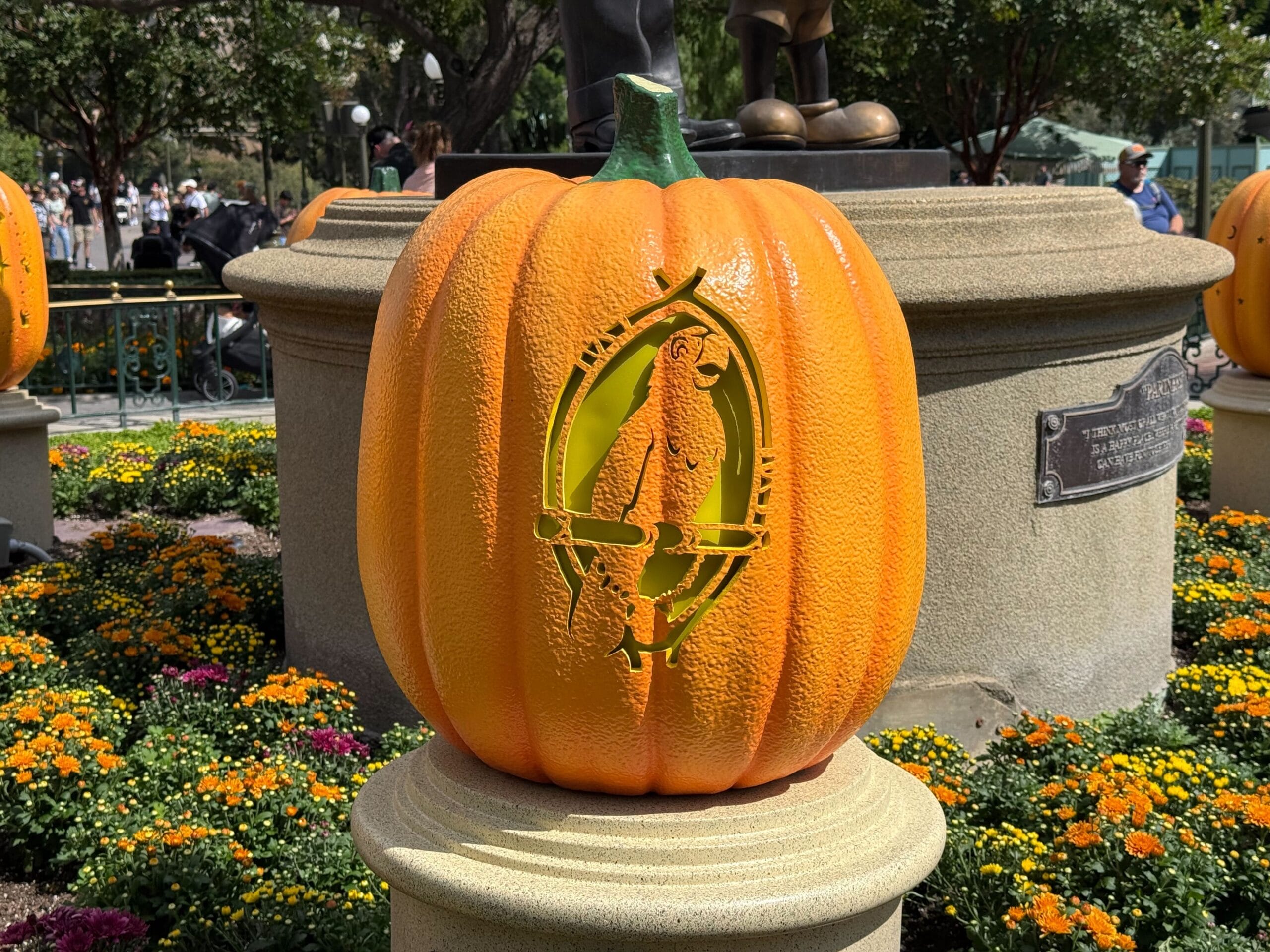 Large orange pumpkin with a carved parrot sitting on a perch, displayed outdoors among yellow and orange flowers.