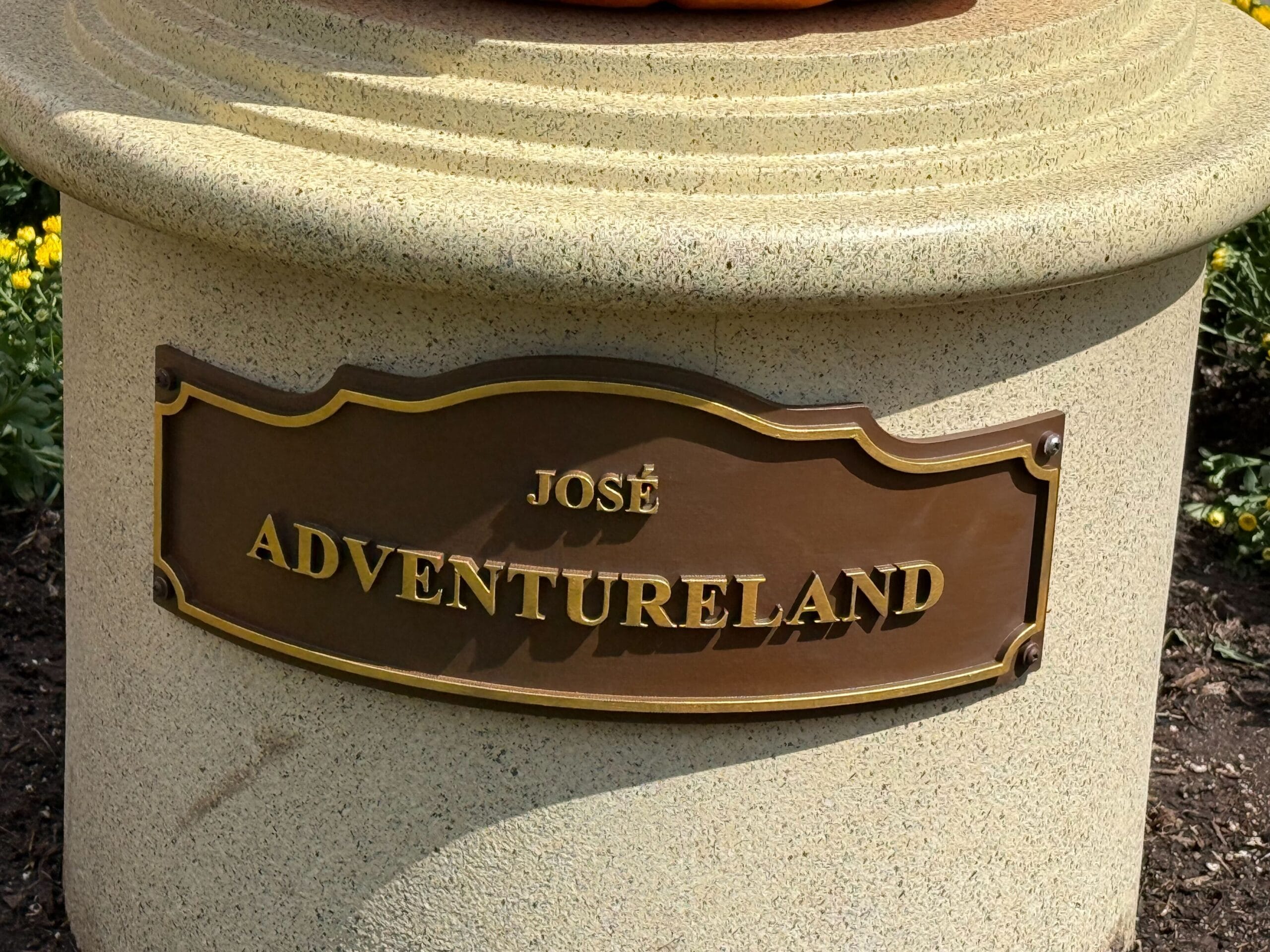 A bronze plaque reads "José Adventureland" on a stone pedestal, with yellow flowers in the background.