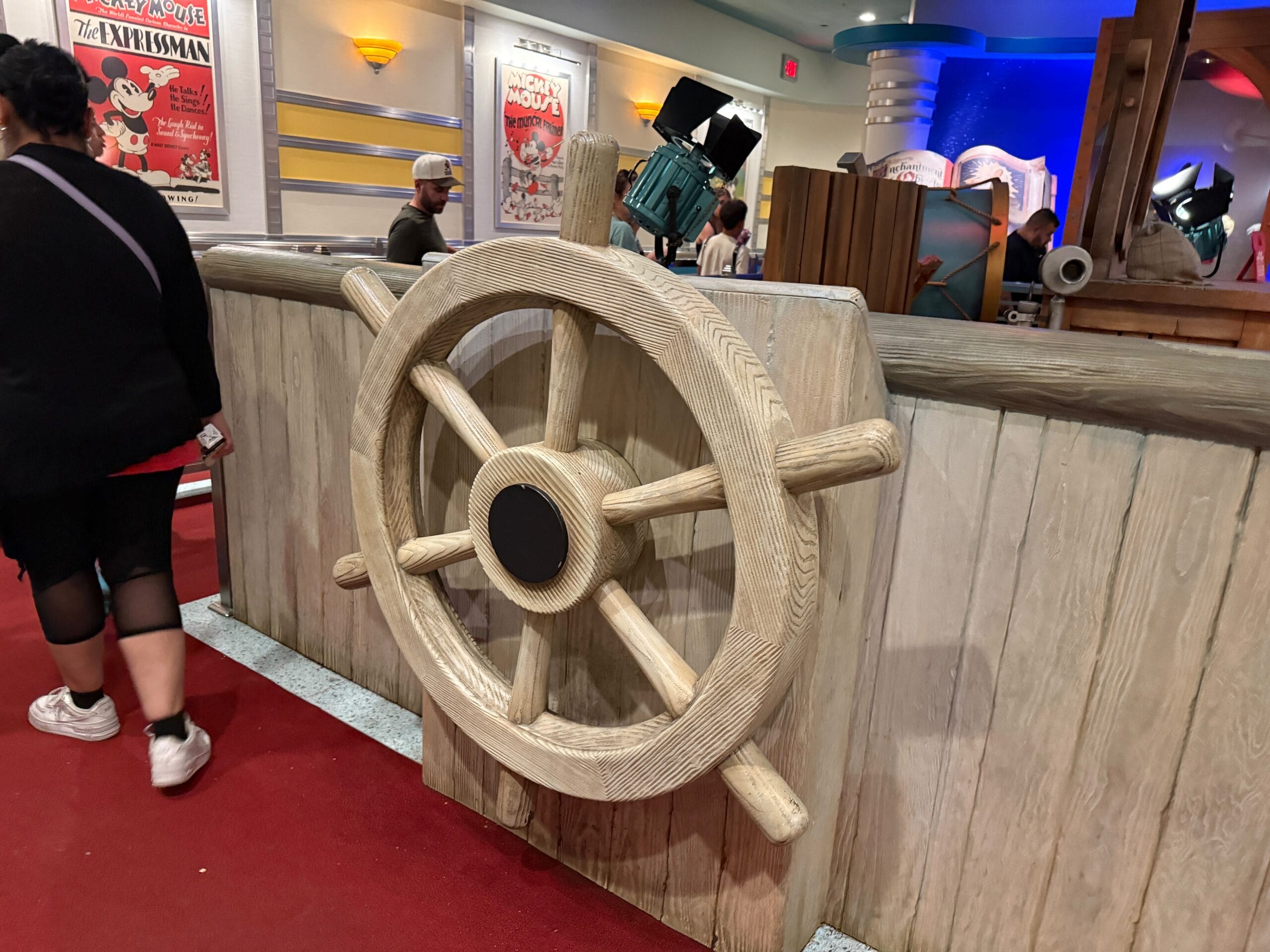 Large wooden ship wheel on a wall indoors, with Mickey decor and people mingling in the colorful background.