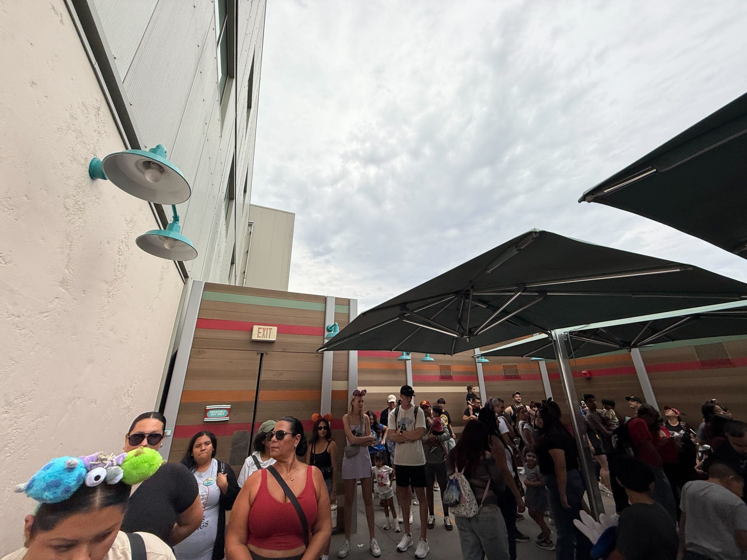 People wait in an outdoor queue under umbrellas, some on cell phones, for Mickey & Minnie's Runaway Railway on a cloudy day.