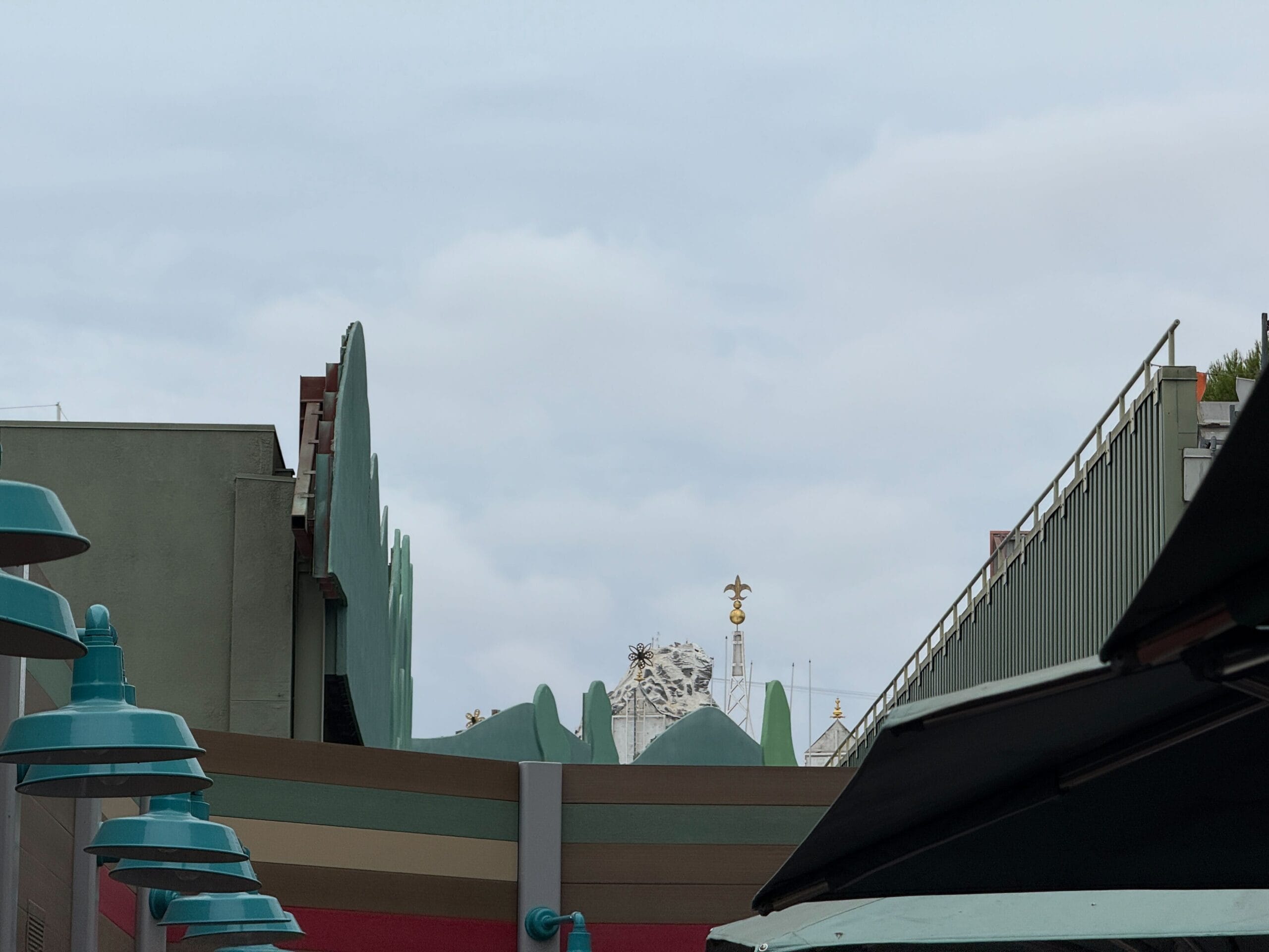 A snowy mountain peak rises above a cityscape as visitors in an outdoor queue use cell phones beneath teal lamp posts.