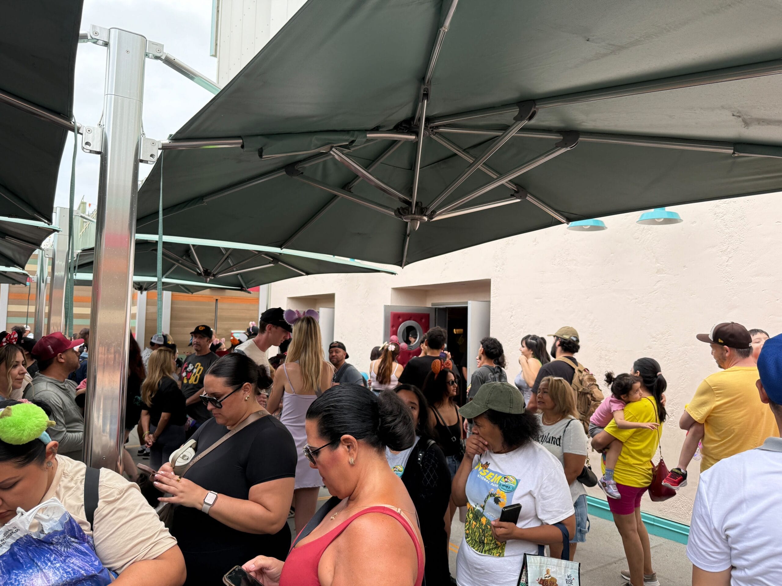 A large outdoor queue waits under umbrellas outside a building, many engaged in cell phone use on a cloudy day.