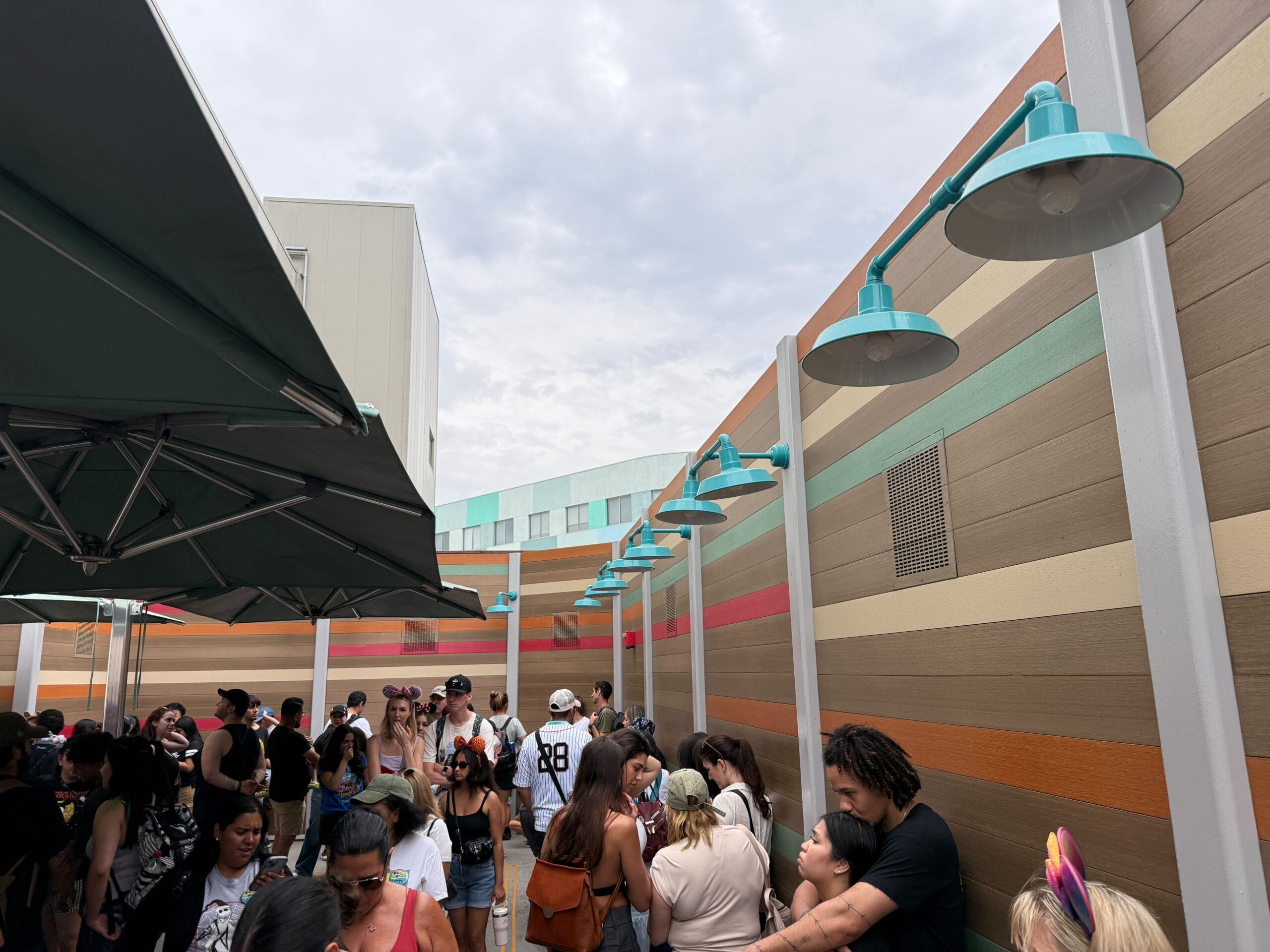 A crowd waits in an outdoor queue beside a colorful striped wall, some engaged in cell phone use under cloudy skies.