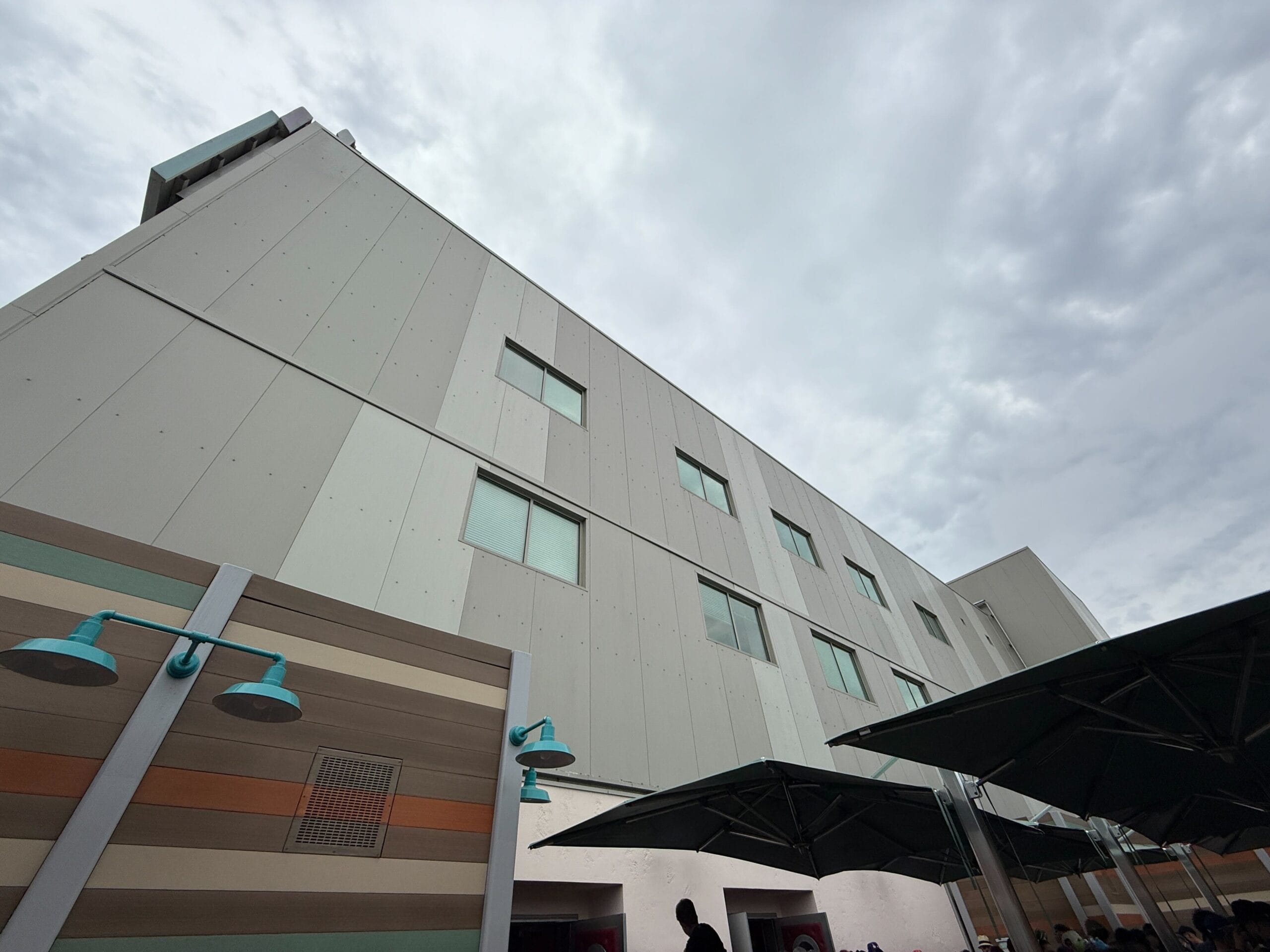 A tall gray building with windows, outdoor lamps, umbrellas, and an outdoor queue under a cloudy sky.