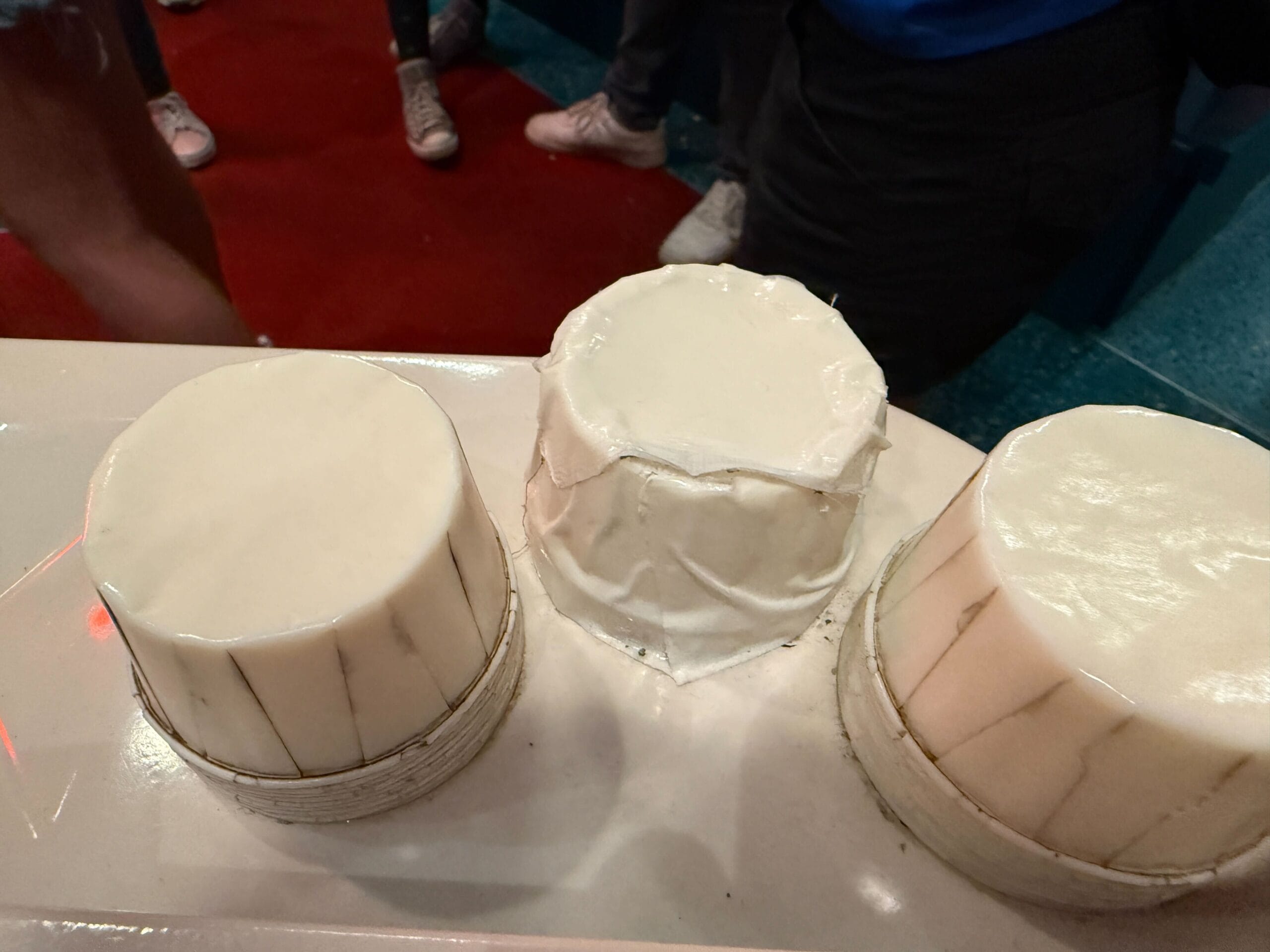 Three round, smooth, white cakes are displayed on a counter near people in an outdoor queue.