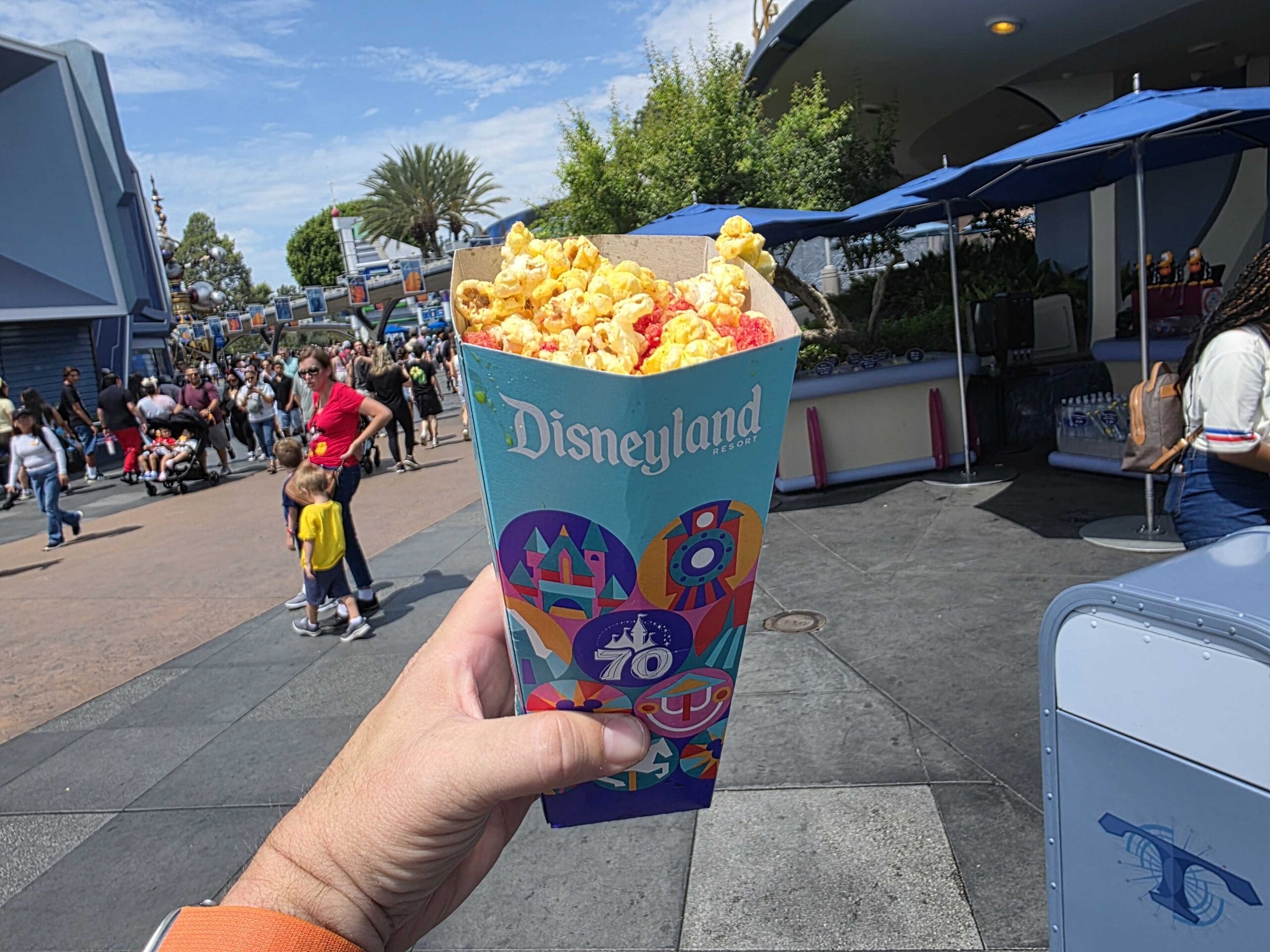 A hand holds a colorful DL Halloween popcorn box outside in a busy park area under a sunny sky.