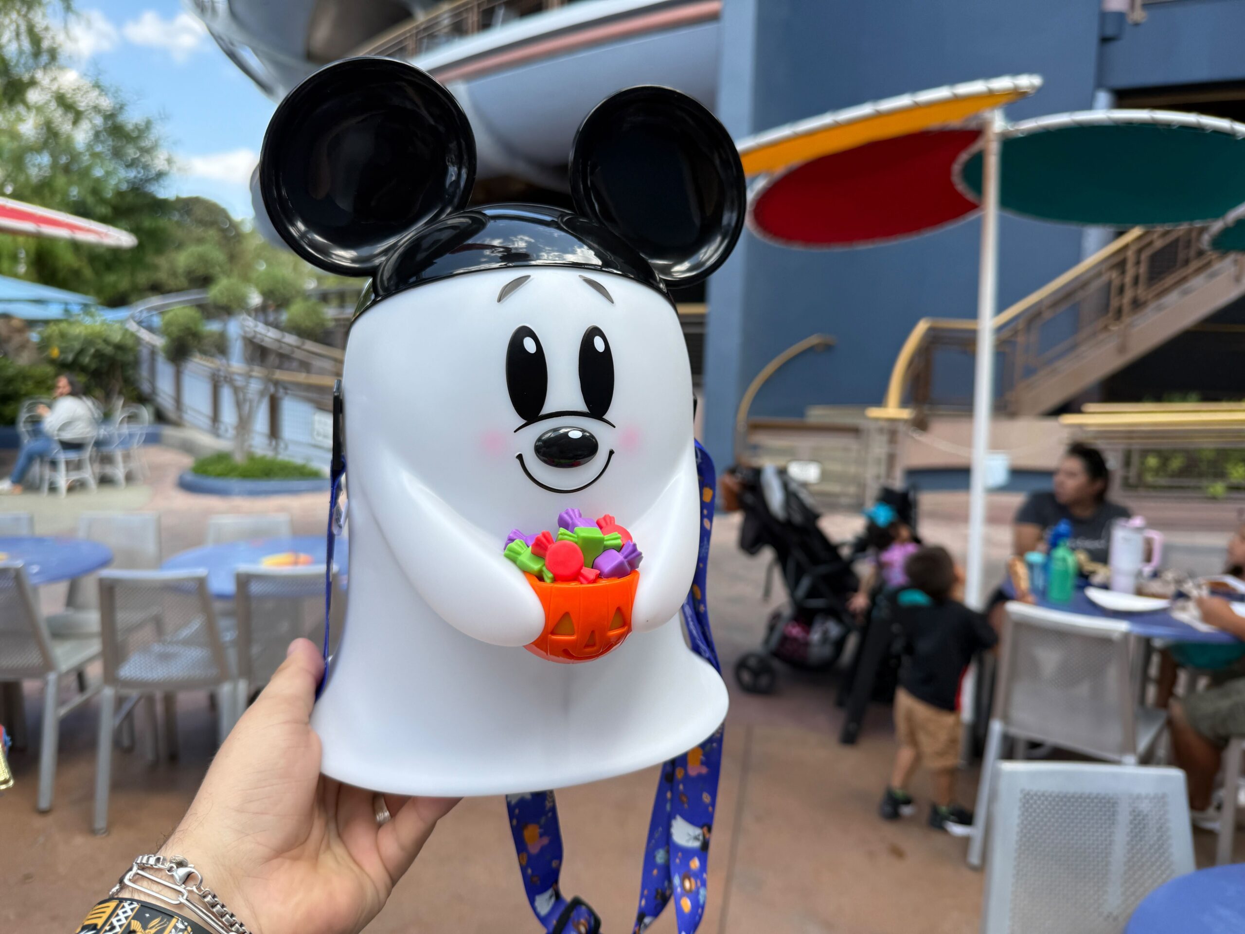 A person holds a Mickey Ghost Popcorn Bucket with a Halloween theme at an outdoor Disneyland Resort dining area.