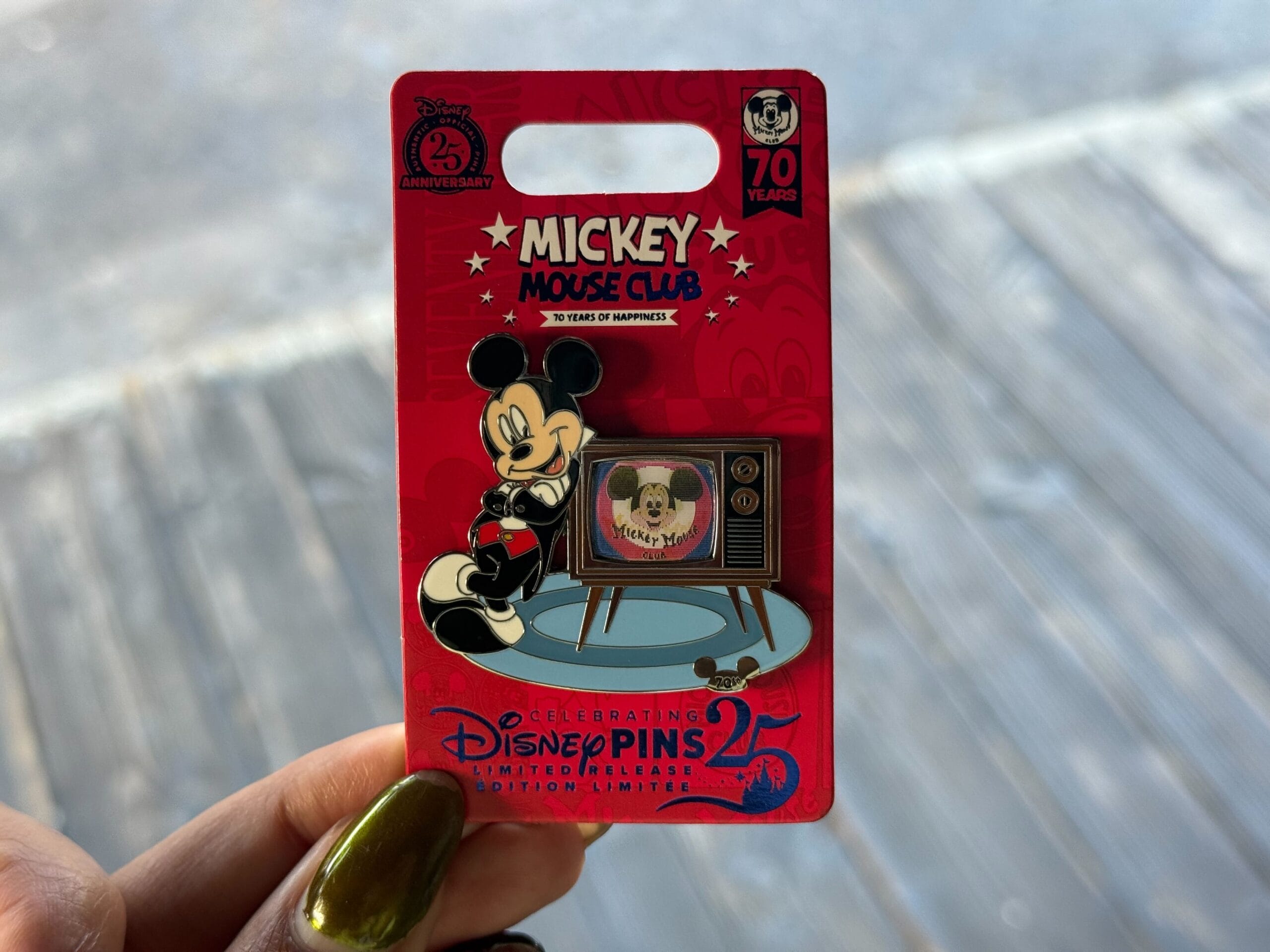 A hand holds a Disney pin featuring Mickey Mouse and a TV, celebrating 70 years of the Mickey Mouse Club.
