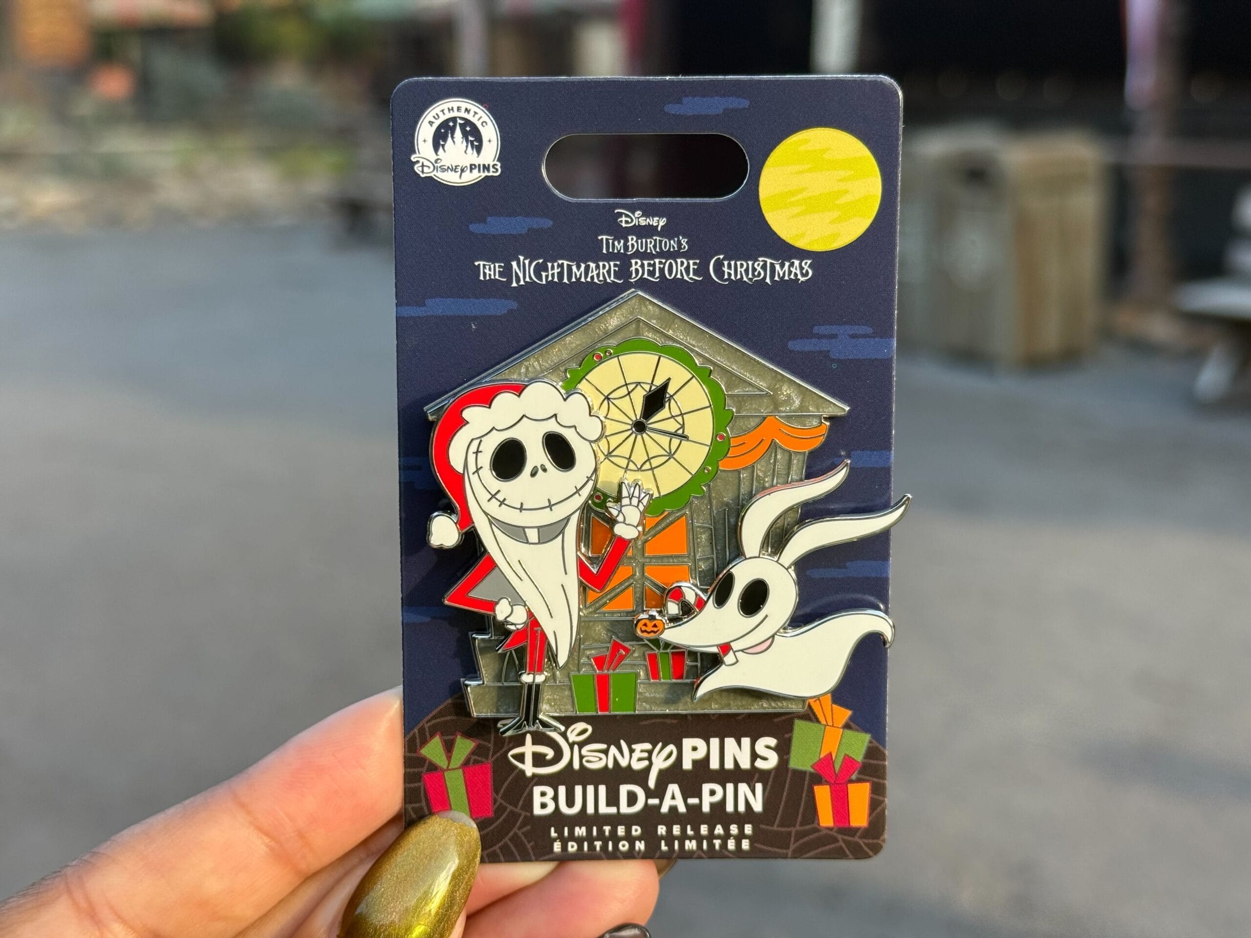 Hand holding a Nightmare Before Christmas Disney pin featuring Jack Skellington and Zero in front of a decorated house.